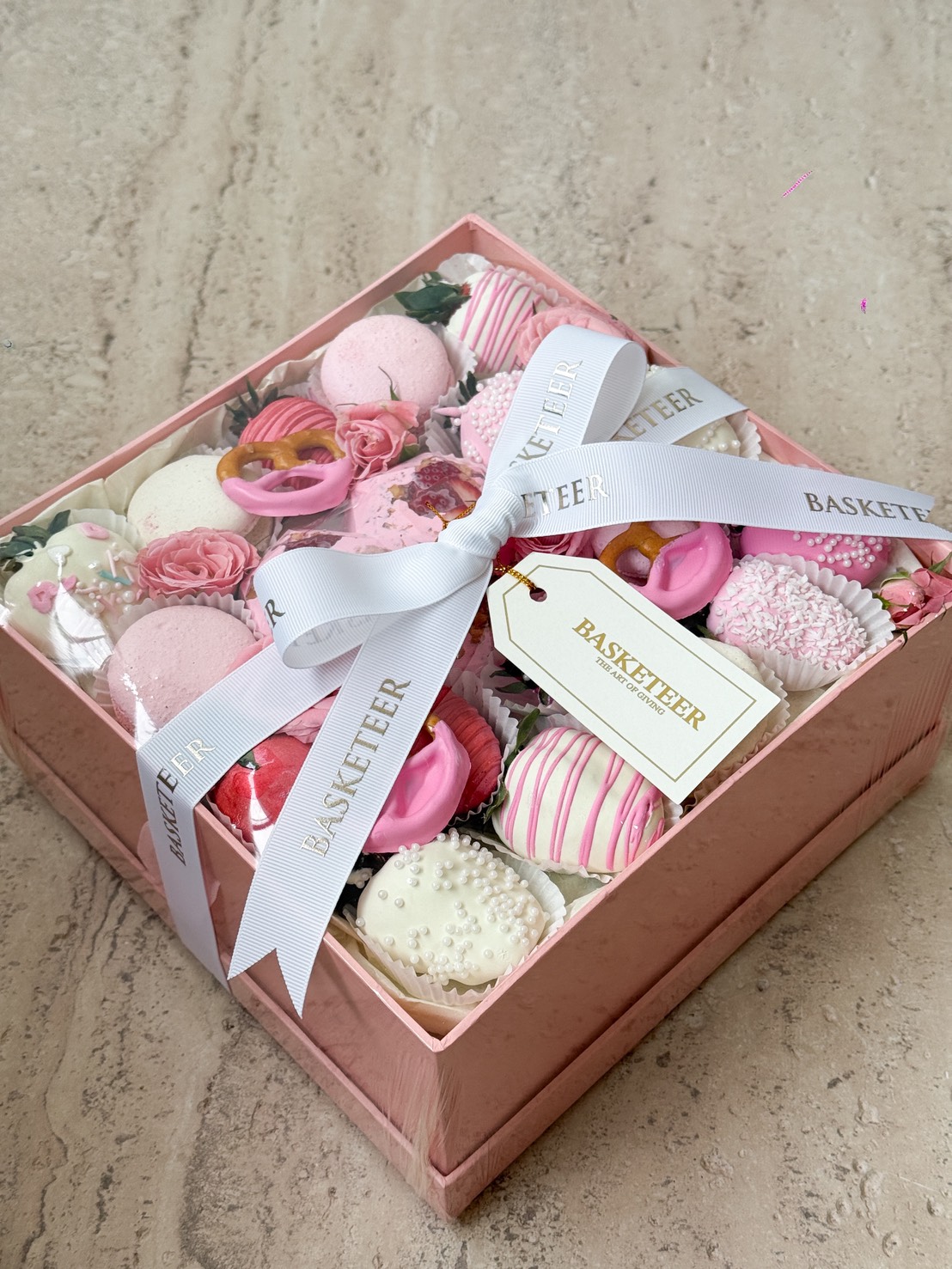 Elegant pink dessert gift box filled with pink and white sweets, macarons, chocolate pretzels, and roses, tied with white Basketeer ribbon.