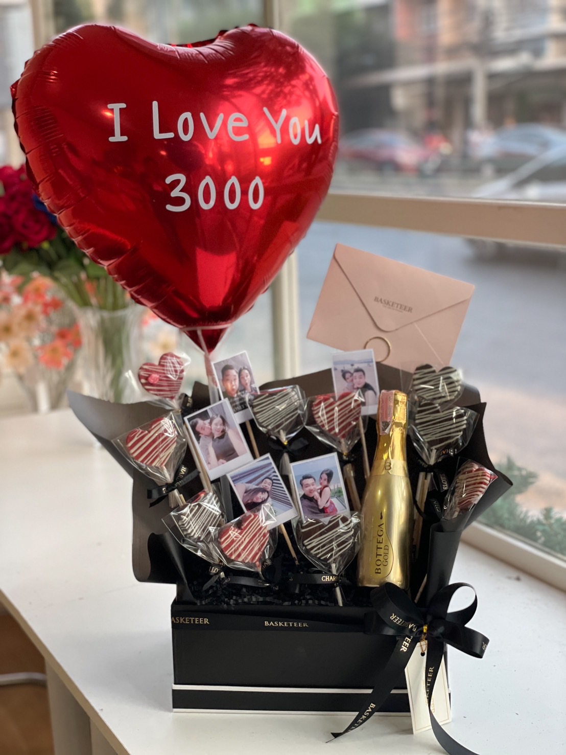 A romantic gift box with heart-shaped chocolates, personalized photo sticks, a gold Bottega champagne bottle, and a red heart balloon reading "I Love You 3000", elegantly wrapped in black.