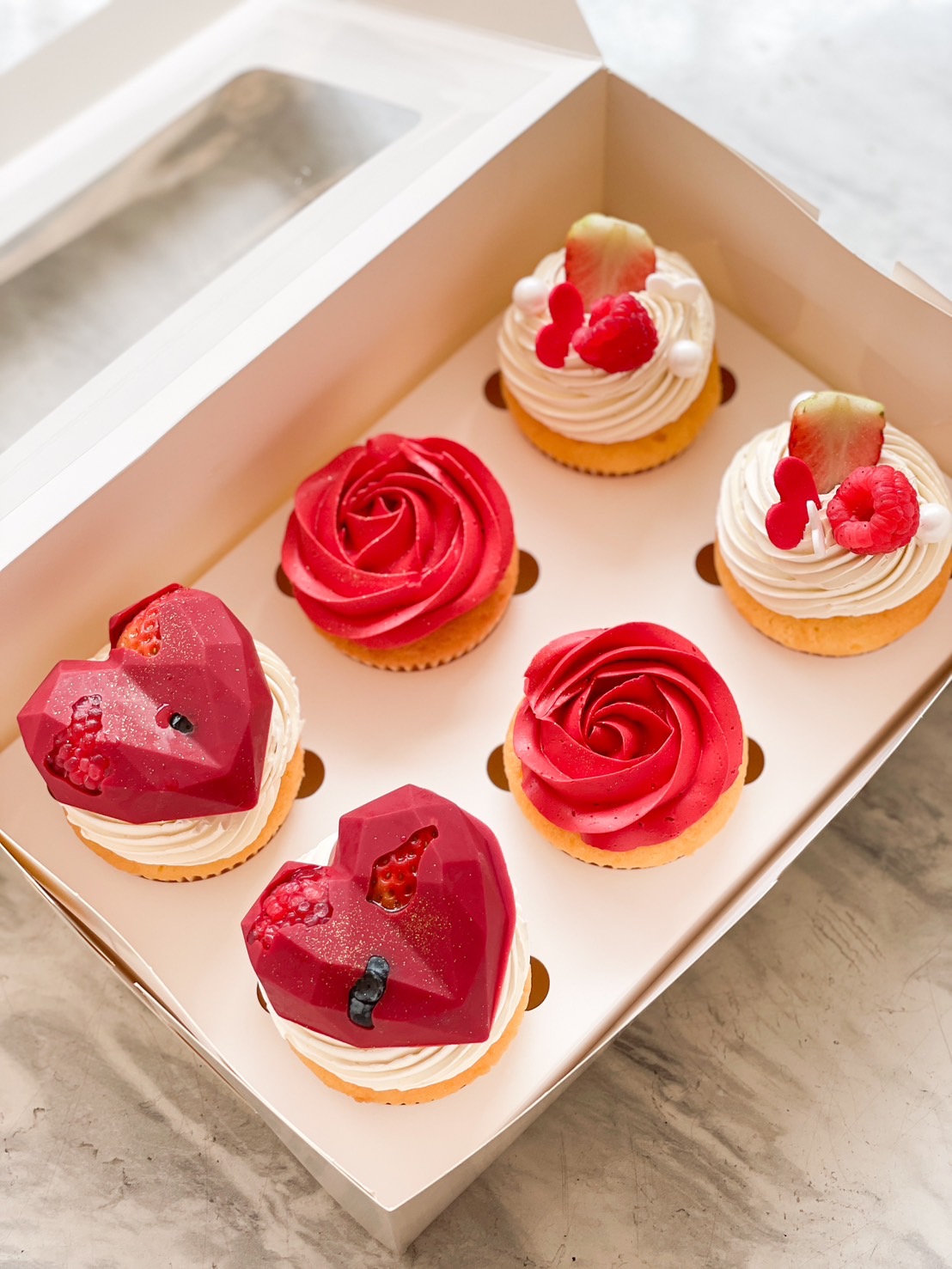 A box of Valentine’s-themed cupcakes, featuring heart-shaped chocolate cupcakes, red rose buttercream swirls, and berry-topped cupcakes, elegantly presented for a romantic surprise.