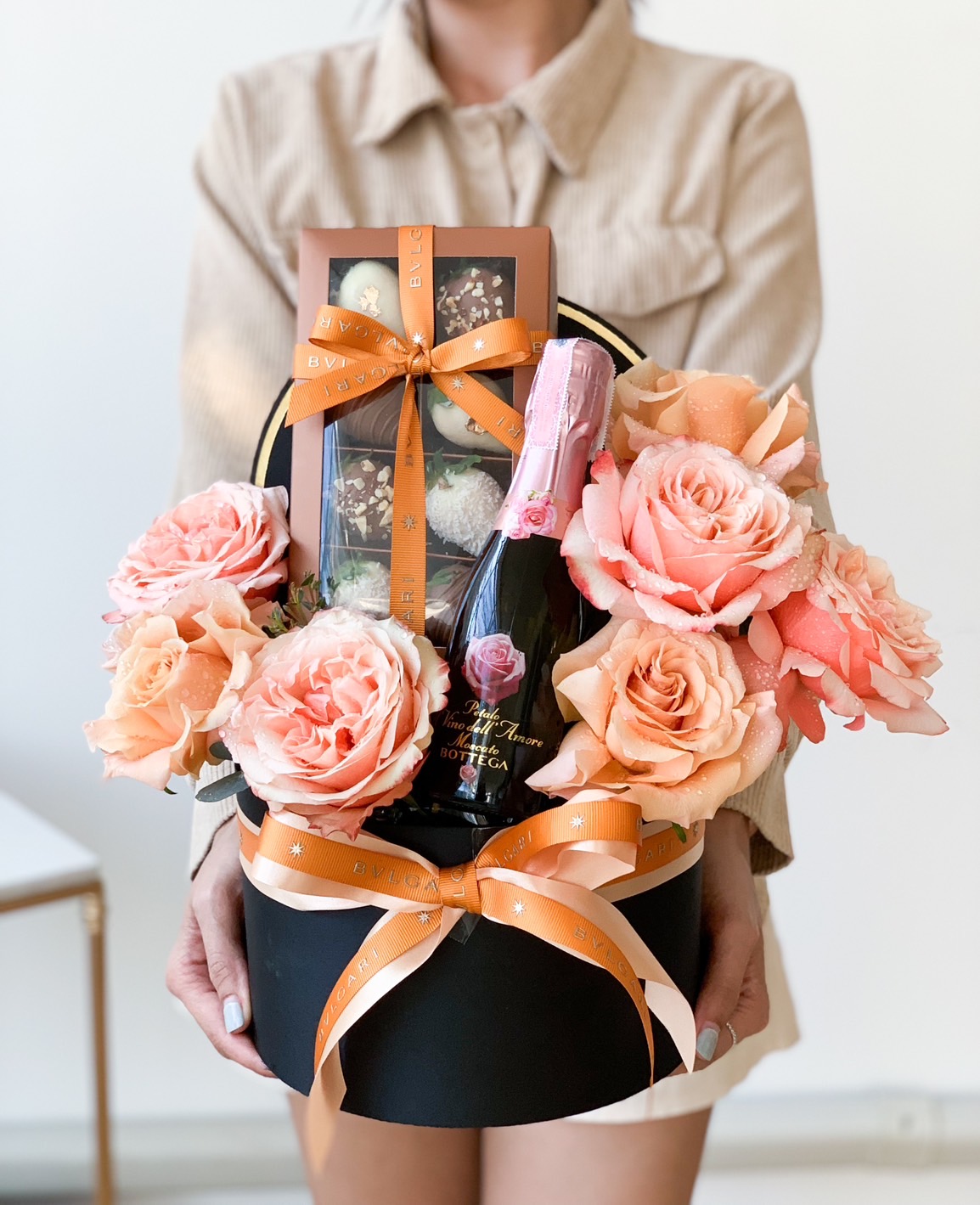 A person in a beige outfit is holding a black hatbox filled with peach roses, a pink sparkling wine bottle, and a box of chocolates. The container is adorned with orange ribbons. A small table can be seen in the background.