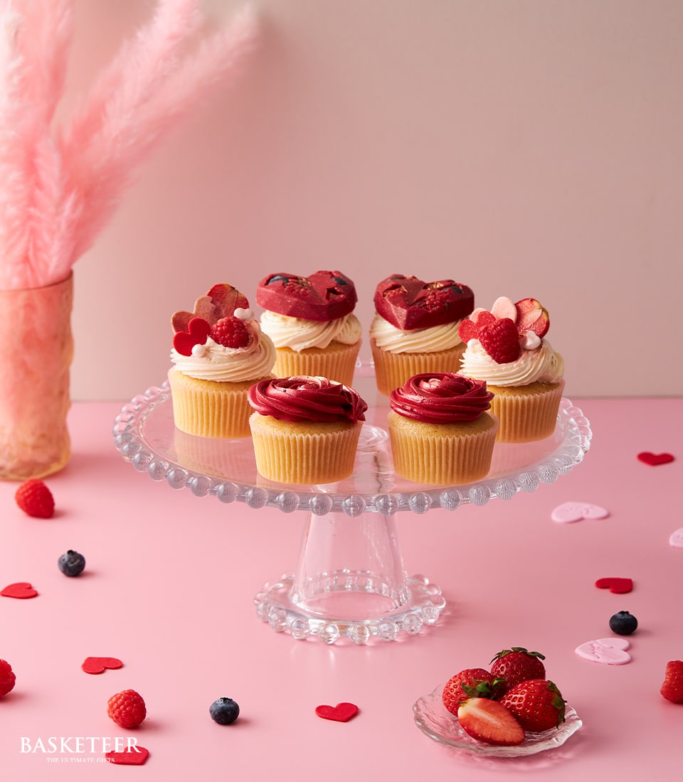 A magnificent glass cake stand elegantly displays the Basketeer Fall In Love Cupcake Set, showcasing seven exquisitely decorated cupcakes adorned with rich red and pristine white icing, heart-shaped embellishments, and succulent fresh berries. In the background, a stylish vase filled with soft pink pampas grass adds a touch of sophistication. Surrounding the stand on the plush pink surface are scattered strawberries, blackberries, and charming pink heart cutouts, creating an inviting and romantic scene. The prestigious Basketeer logo adorns a ribbon encircling one of the treats, affirming its luxurious presentation.