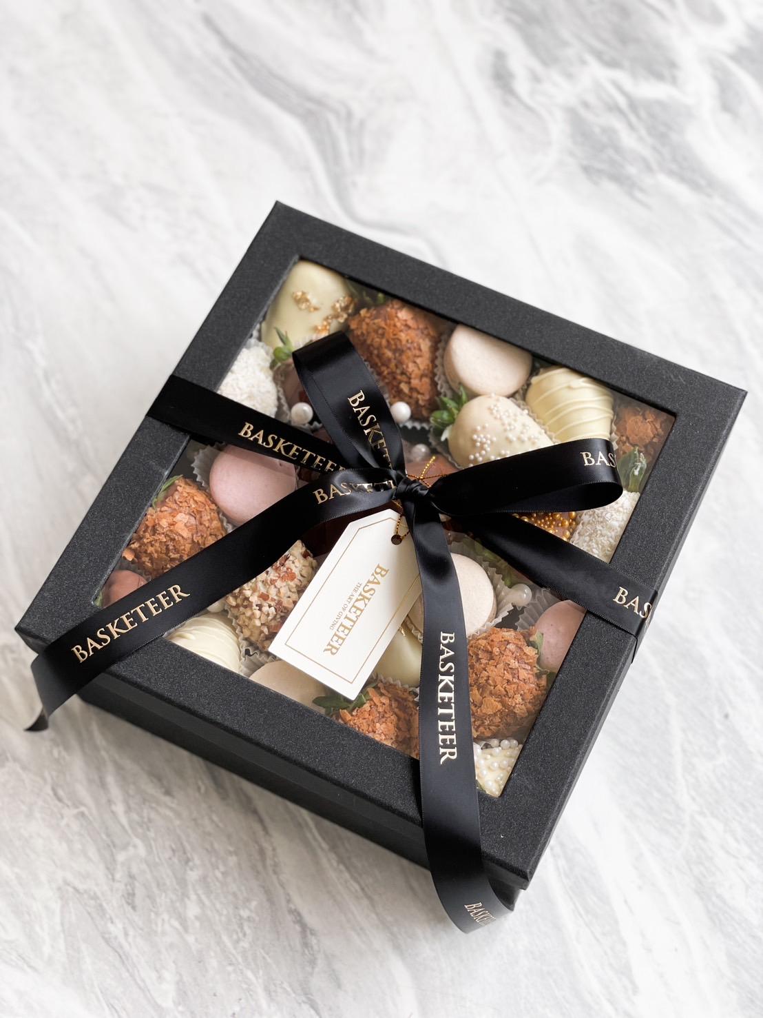 A black gift box with a transparent lid showcasing assorted confections, including a heart chocolate & cherry gift set, wrapped with a black ribbon labeled 