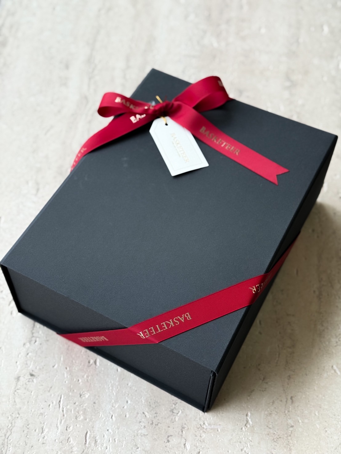 Matte black luxury gift box tied with red satin BASKETEER ribbon and white tag