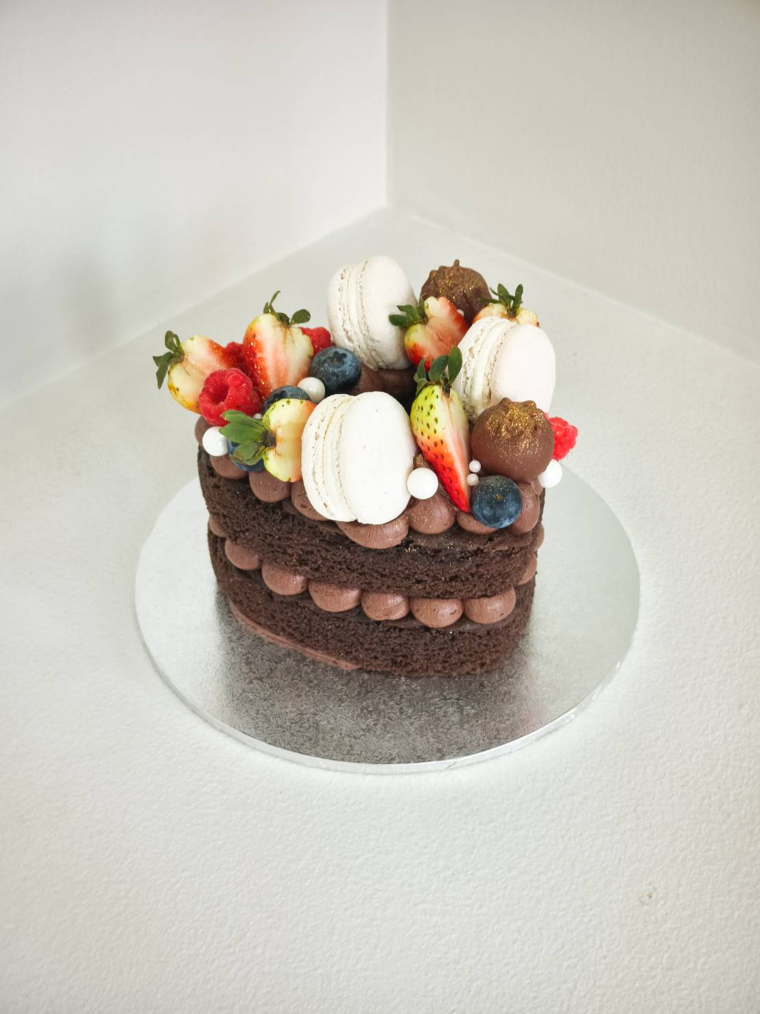 A heart-shaped Charlotte chocolate cake adorned with macarons, fresh berries, and pearls on a silver board, baked in Bangkok.