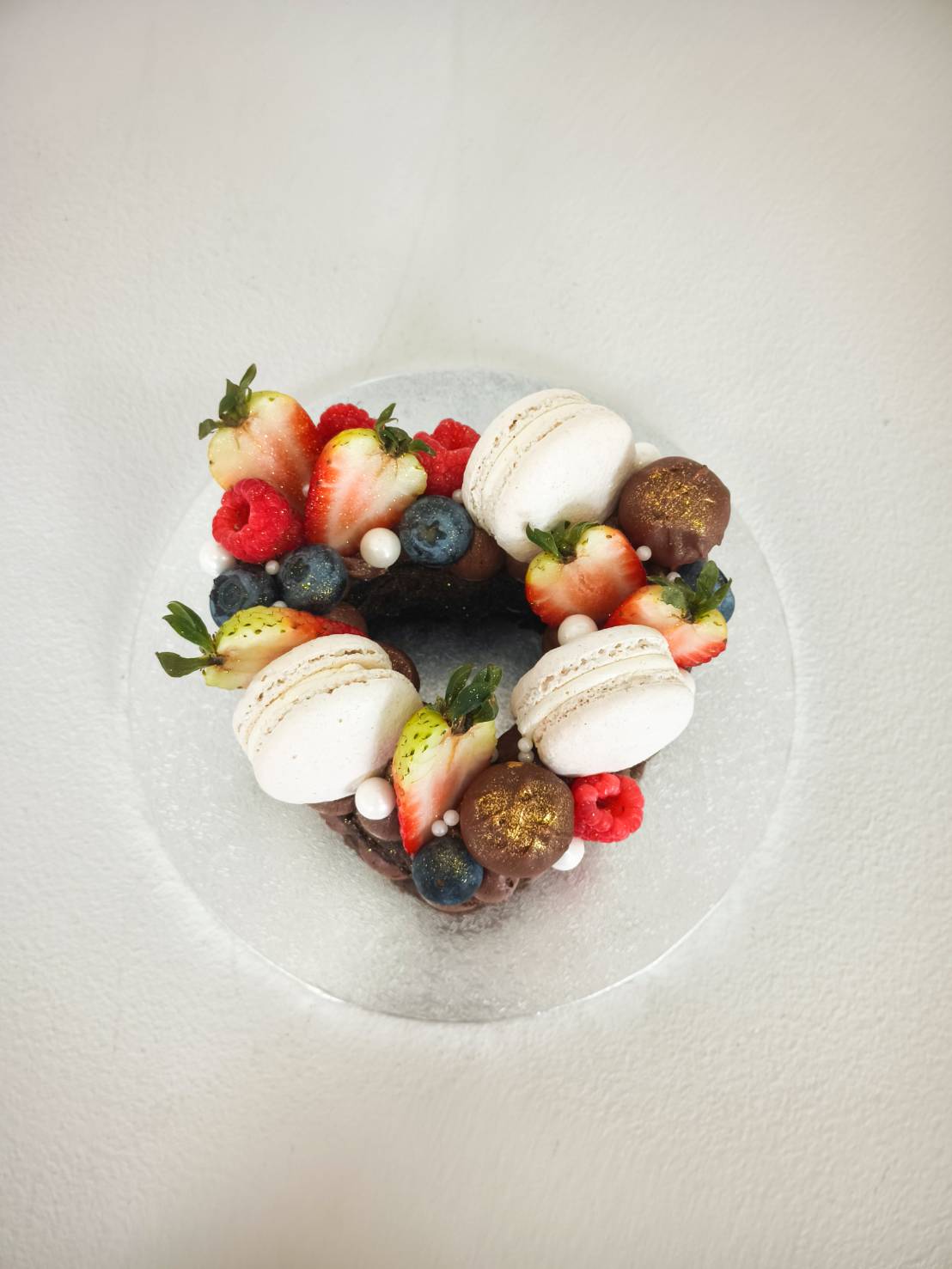 A heart-shaped Charlotte cake crowned with white macarons, fresh berries, chocolate truffles, and meringues on a glass platter.