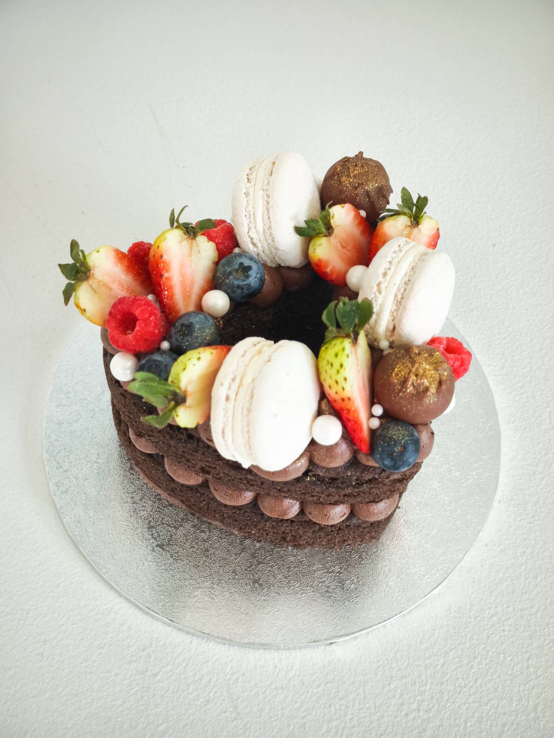 A heart-shaped Charlotte chocolate cake adorned with macarons, fresh berries, chocolate-dipped strawberries, and sugar pearls.