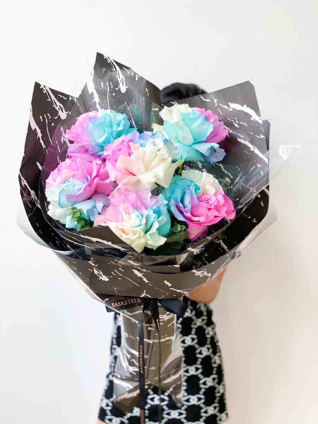 A person holds a large bouquet of multicolored roses wrapped in black and white marbled paper with a black ribbon. The roses are in shades of pink, blue, white, and light green. The person’s face is obscured by the bouquet.
