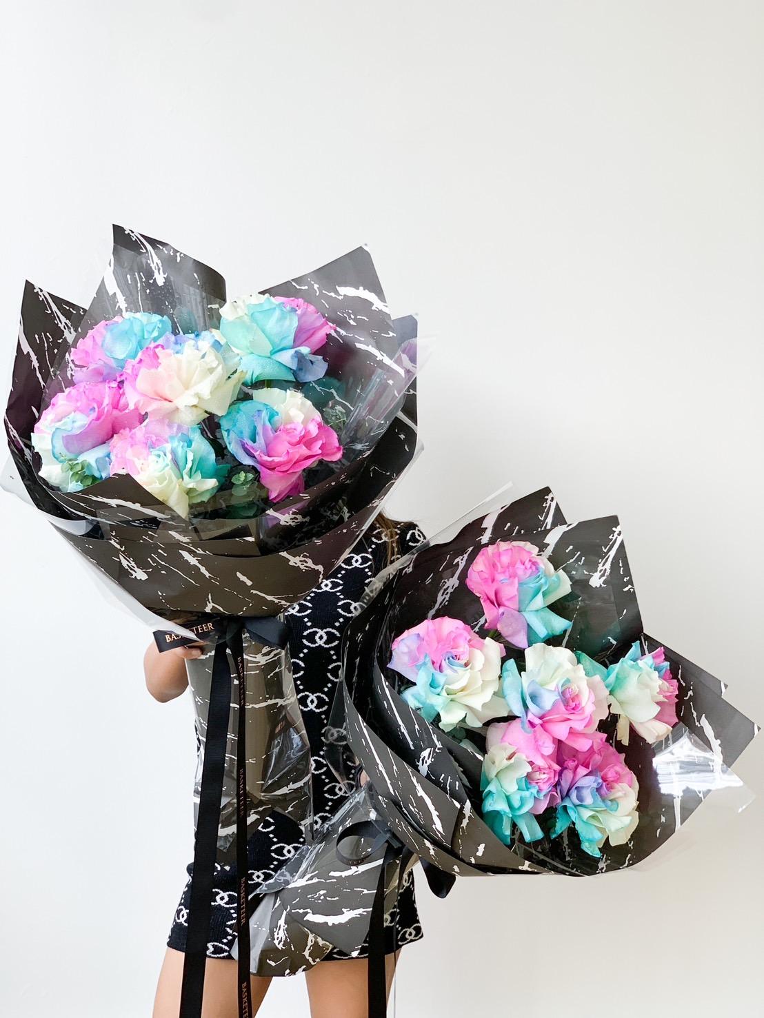Two bouquets of roses, each featuring a mix of pastel colors including blue, pink, and white, wrapped in black and white marbled paper and tied with black ribbons. Both bouquets are being held by a person wearing a black and white patterned dress against a plain background.