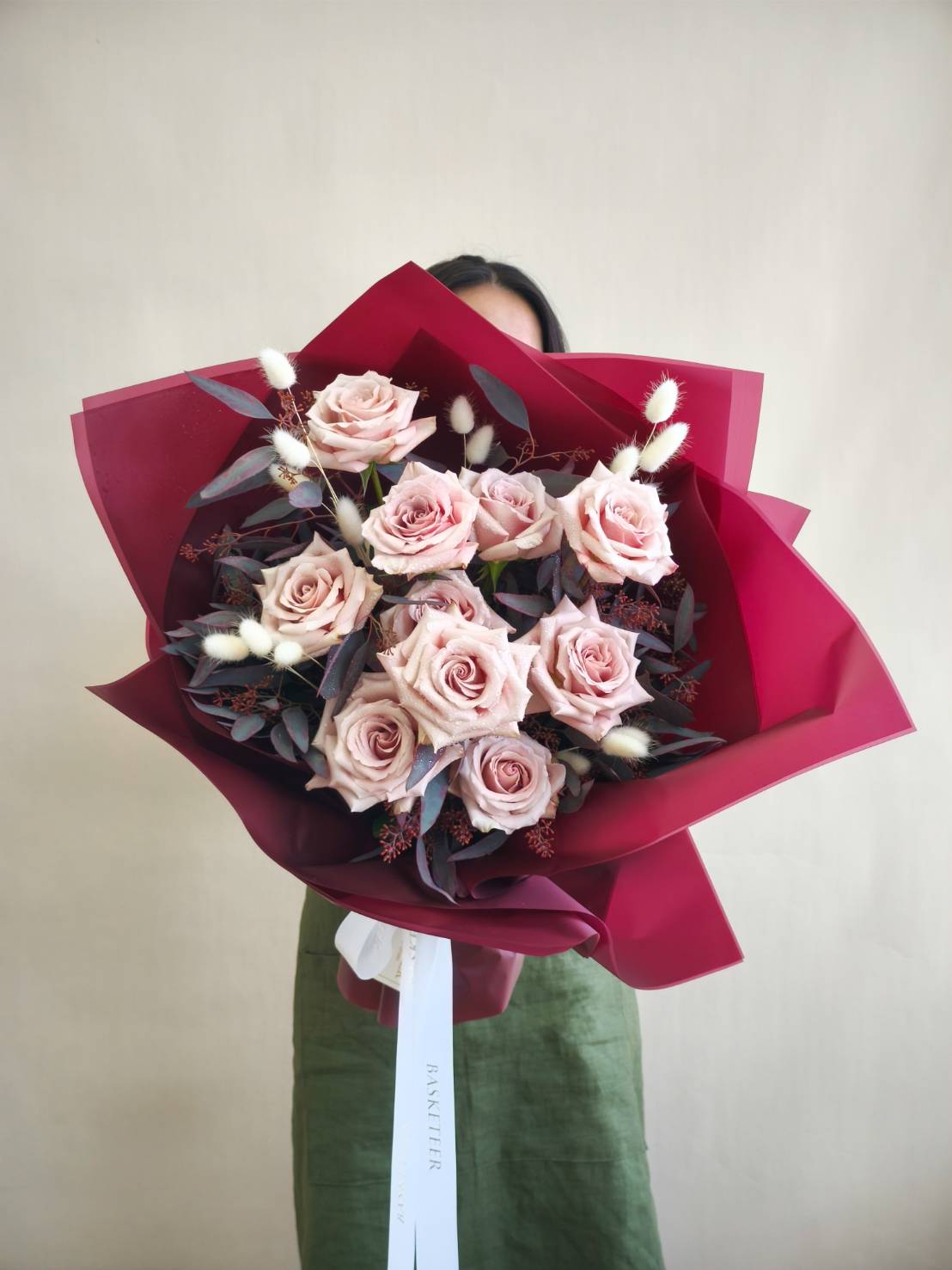 Elegant bouquet of nude pink roses with white bunny tails, wrapped in deep red paper and tied with white ribbon.