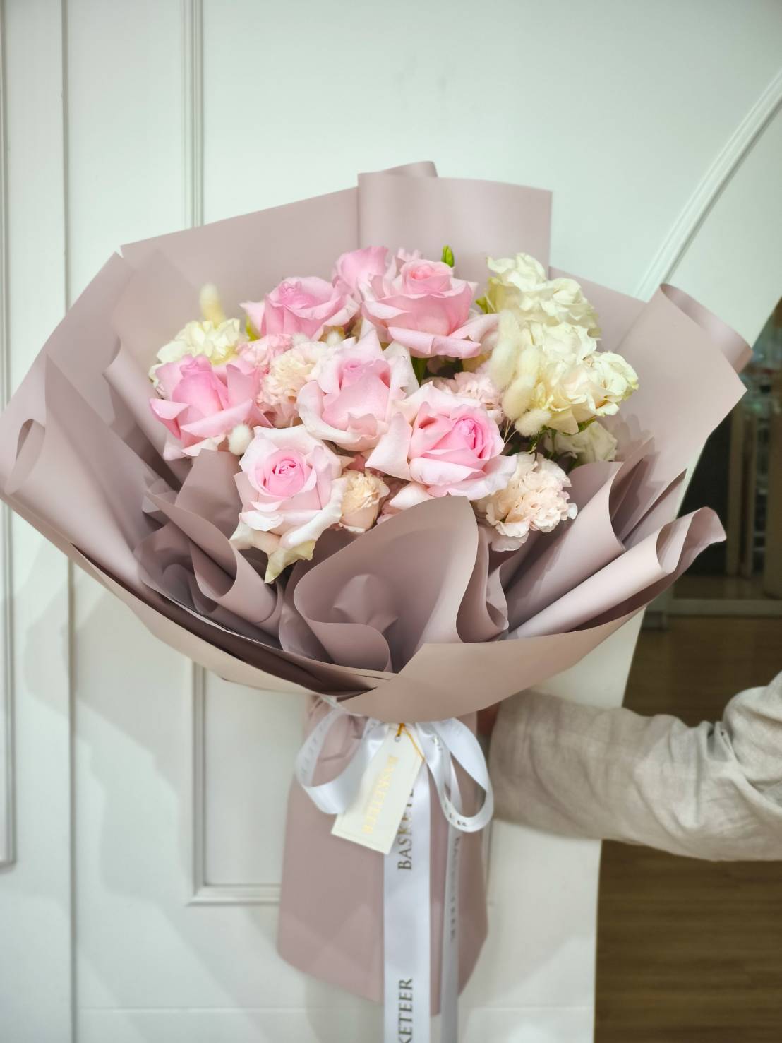 Elegant bouquet of blush pink roses, cream blooms, and pastel flowers wrapped in soft mauve paper with satin ribbons.