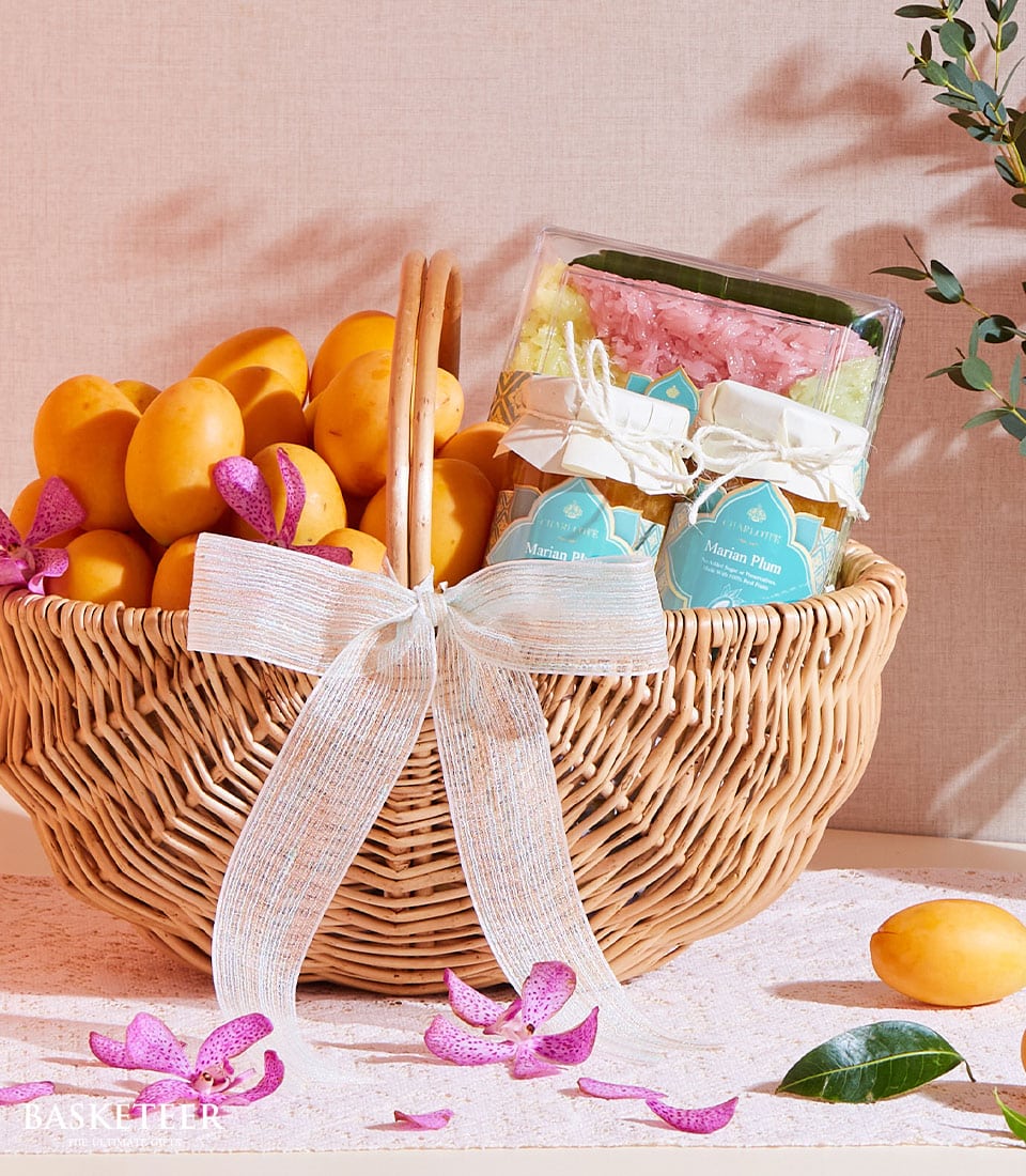 A luxurious wicker basket teeming with vibrant, sun-kissed yellow mangoes and two elegantly packaged dried marian plums is exquisitely curated as the Marian Plum & Sticky Rice Gift by Basketeer. The basket is adorned with a pristine white ribbon featuring the Basketeer logo, embodying refined elegance. Delicate pink flower petals are artfully scattered around the display, with a solitary mango placed just outside the basket for an inviting touch. In the upper right corner, a lush leafy branch adds a natural sophistication to this sumptuous gift presentation.