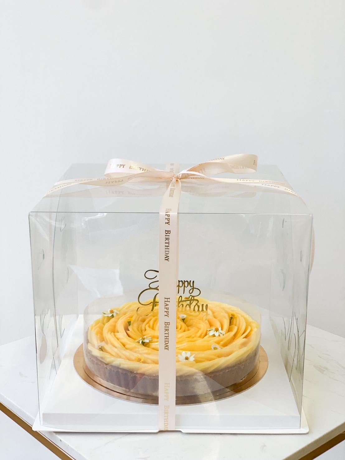 A beautifully decorated Namdokmai Mango Cheese Tart with rose-like frosting is enclosed in a clear, square plastic box. The box is tied with a light-colored ribbon that reads 