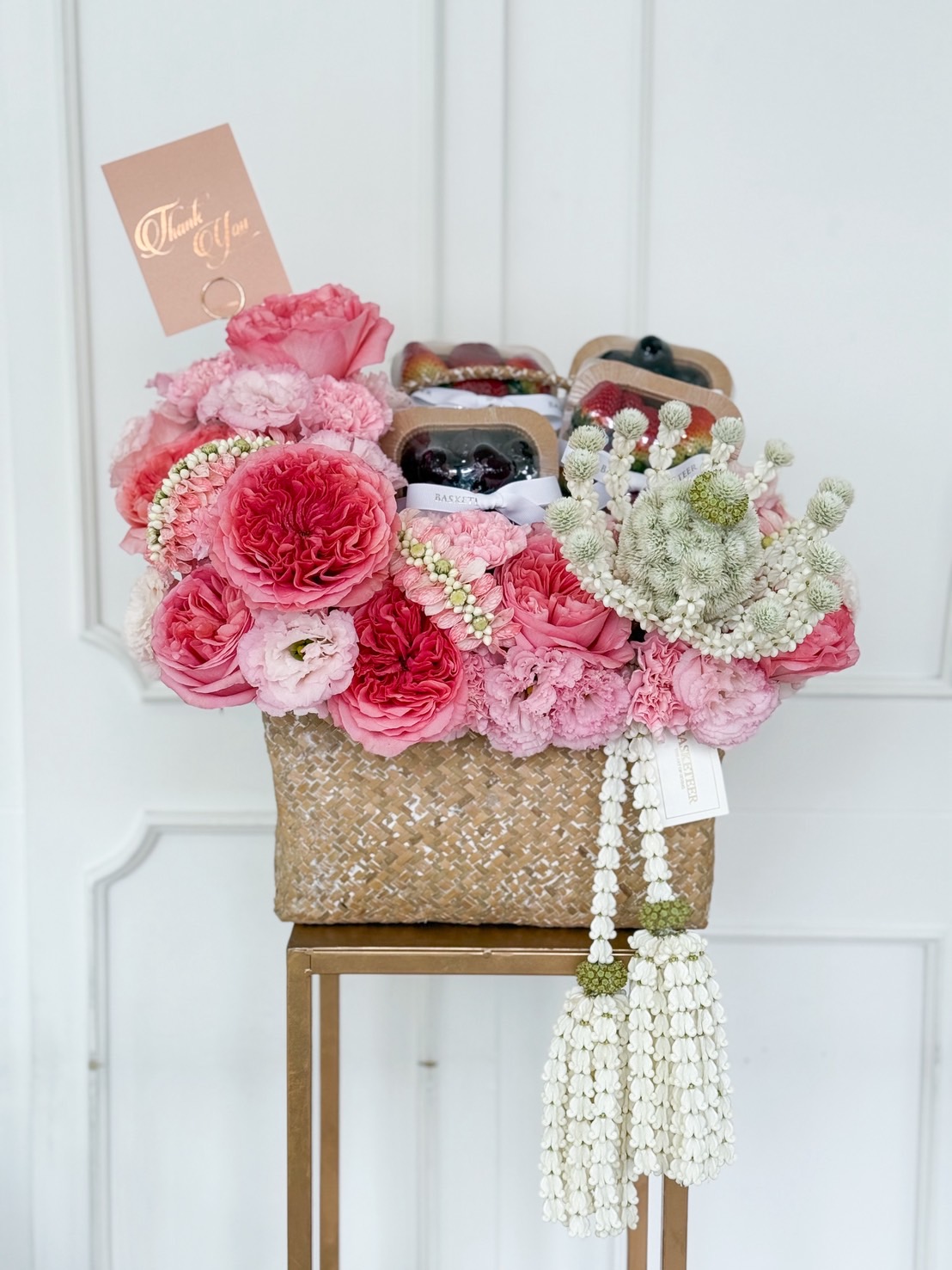Luxury woven basket with pink roses, carnations, and peonies, paired with premium fresh fruits and a traditional Thai jasmine garland.