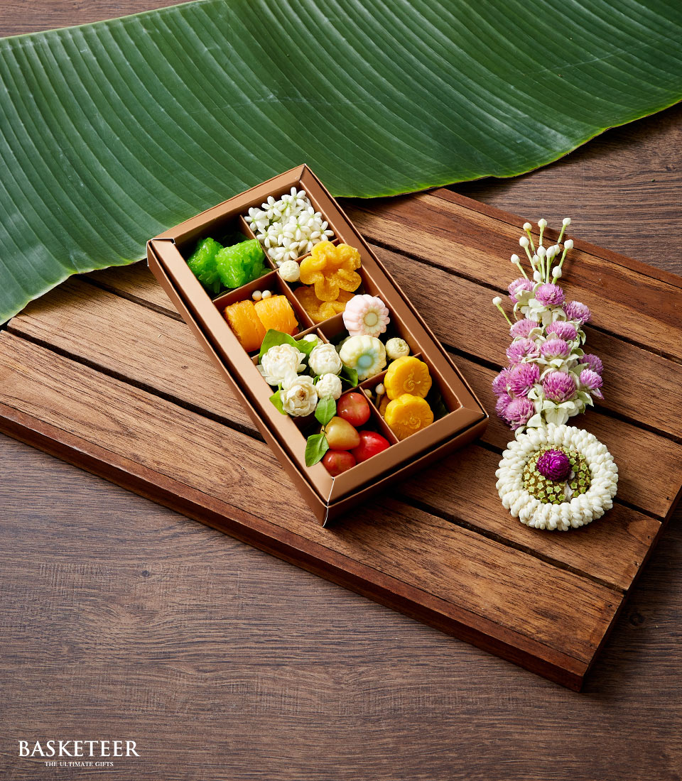 A meticulously crafted rectangular box of assorted, vibrant Thai desserts from Basketeer graces a wooden table, elegantly presented atop a lush green leaf. To the side, a delicate garland of white and purple flowers enriches the scene with celebratory flair. The prestigious Basketeer logo is tastefully displayed in the bottom left corner, embodying luxury and sophistication.