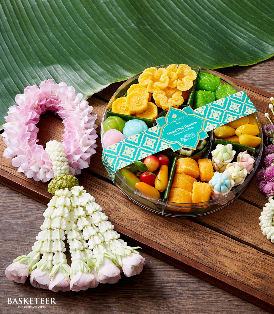 A luxurious round box of Songkran Sweets by Basketeer is elegantly presented on a rich wooden surface, adorned with vibrant traditional Thai delicacies and a refined green label. Nearby, an exquisitely crafted floral garland in soft pinks and whites, showcasing intricate details, gracefully rests atop a lush green leaf, completing the inviting scene. The Basketeer logo proudly adorns the packaging ribbon, encapsulating the essence of opulence and heritage.