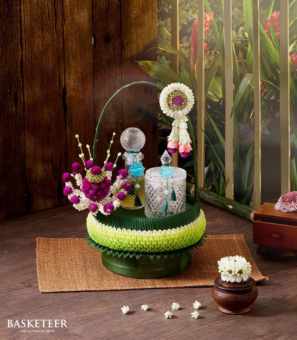 An opulent and exquisitely ornate floral arrangement graces a polished small table, emanating luxury and charm. The centerpiece, adorned with luscious white, delicate pink, and regal purple blooms, harmoniously complements the crystal glassware that sparkles under soft lighting. Lavish intricate floral decorations further elevate the scene. To add an exotic touch, a traditional Songkran Thai incense set is thoughtfully placed within the arrangement. The sophisticated setting is enriched by the backdrop of rich wooden paneling and a serene view of lush greenery through expansive windows. A luxurious box tied with a ribbon bearing the distinguished Basketeer logo subtly suggests refinement and attention to detail in every gesture.