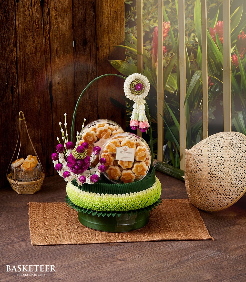 A luxurious food gift set by Basketeer, showcasing two exquisite trays of cookies adorned with intricate floral decorations. The cookies are elegantly placed on a sumptuous green pedestal against a rich wooden backdrop. Enhancing the scene is a tall decorative stand and a beautifully crafted natural woven item, all resting atop a branded mat that proudly reads 