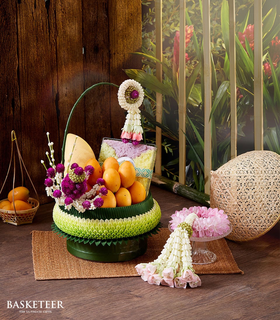 A lavishly decorated basket brimming with vibrant oranges, a luscious pineapple, and exquisite flowers is elegantly adorned with intricate floral garlands. It rests gracefully on a finely woven mat next to another beautifully crafted basket filled with fragrant Thai incense and stunning blossoms. The serene backdrop of rich wooden panels and verdant foliage enhances the inviting ambiance. A tasteful ribbon bearing the distinguished 