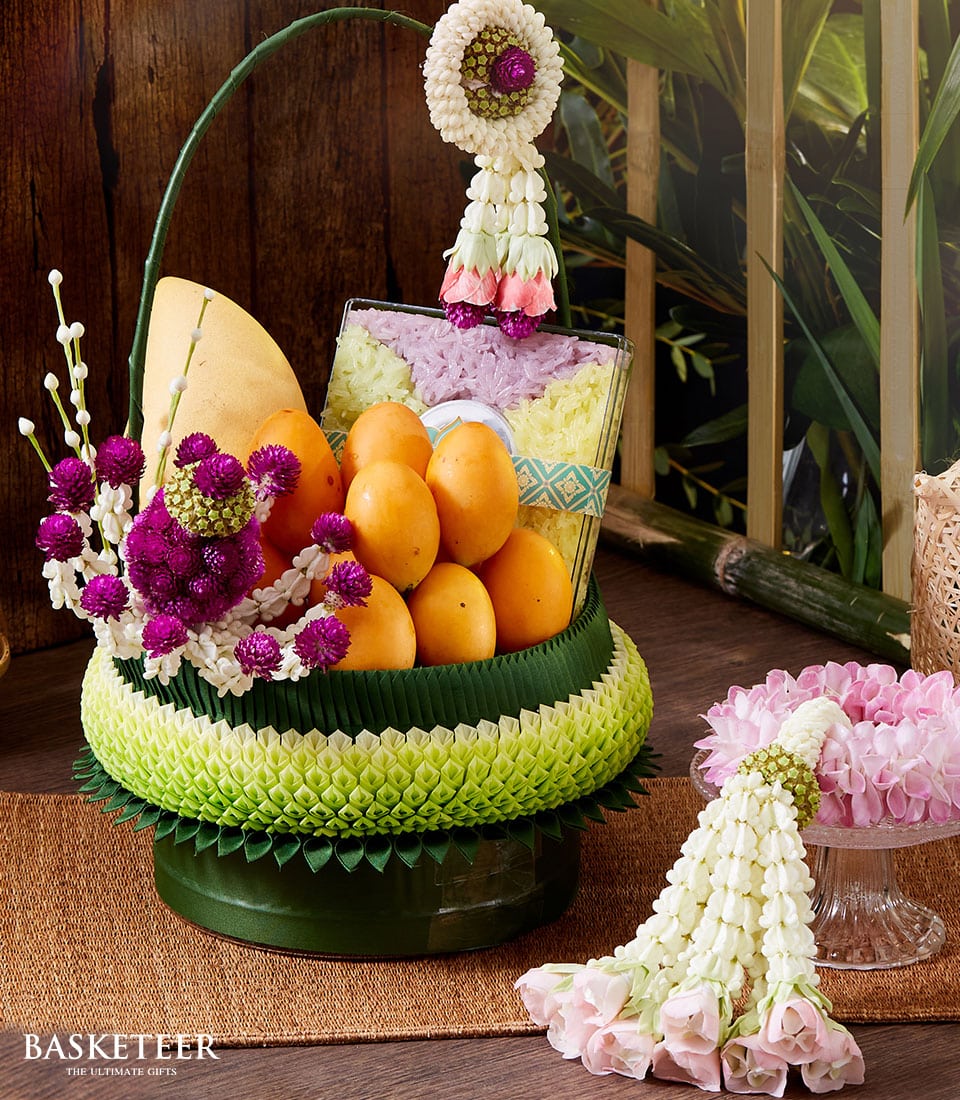 A sumptuous Basketeer fruit basket brimming with luscious, ripe mangoes and intertwined with elegant green decorative elements, beautifully evoking the essence of a Songkran Thai incense set. The arrangement is gracefully adorned with an exquisite garland of white and purple flowers, accompanied by a coordinated smaller floral decoration placed adjacent to the main display. The scene is set against a backdrop of rich wooden elements and vibrant greenery, creating an inviting and luxurious ambiance. A ribbon bearing the Basketeer logo adds a signature touch of sophistication to this stunning presentation.