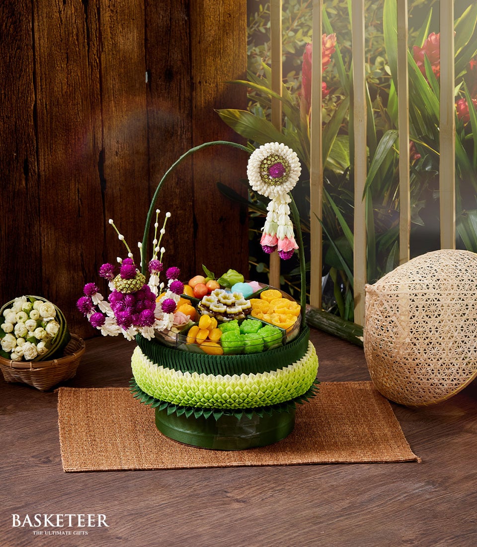 An elegant ensemble of traditional Thai desserts graces a luxurious green pedestal tray, exquisitely adorned with intricate white, green, and purple floral garlands. Behind the tray, a meticulously arranged basket brimming with pristine white flowers and a refined Songkran Thai incense set bears the prestigious Basketeer logo, epitomizing opulence. This lavish display rests on an intricately woven mat set against a lush tropical backdrop, inviting indulgence and serenity.