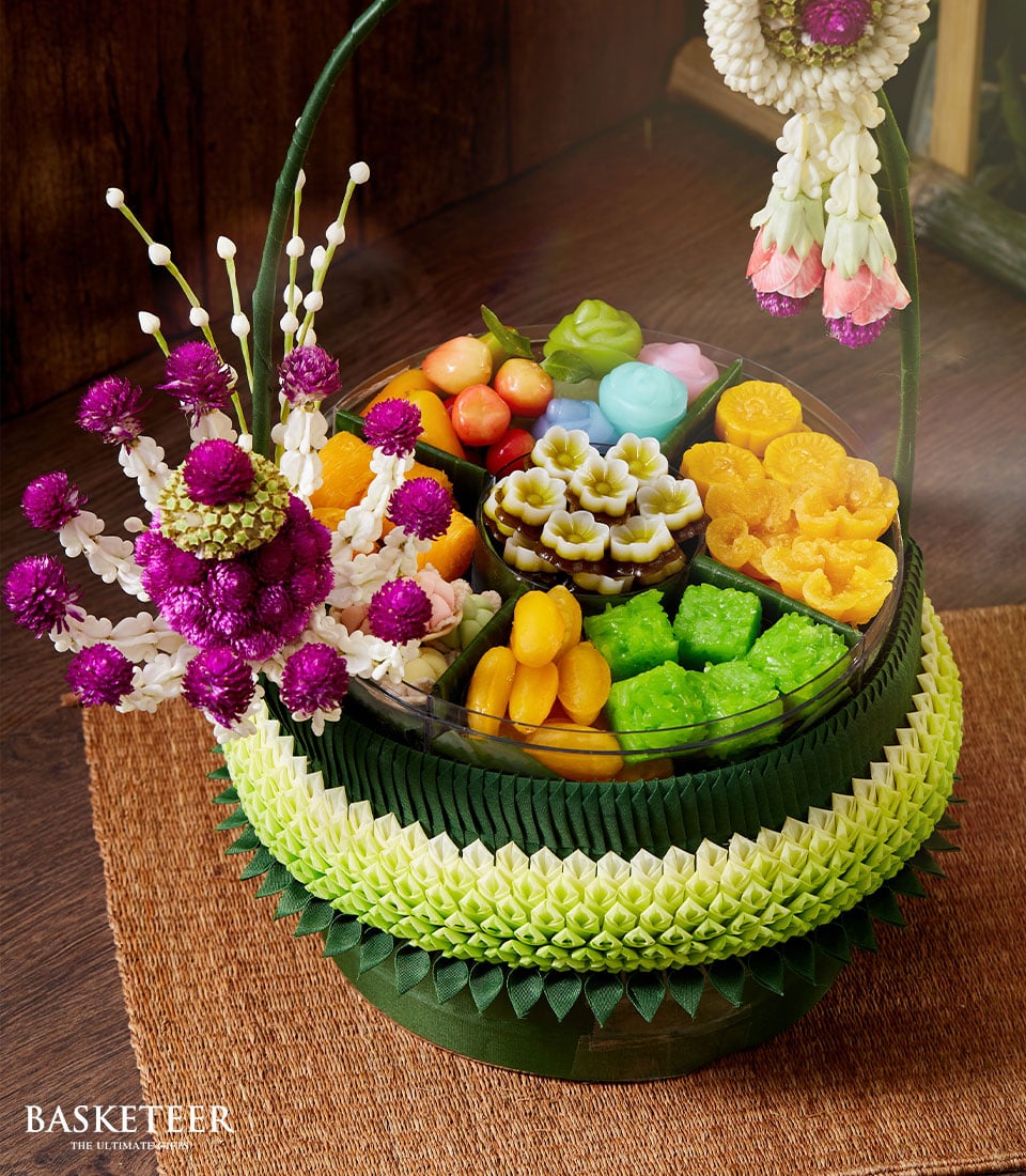 A lavishly arranged Thai dessert basket from Basketeer, showcasing an assortment of vibrant traditional sweets, including exquisite flower-shaped delicacies and elegant layered jelly. The basket is meticulously adorned with green and white folded leaves, embellished with luxurious purple and white blossoms. A ribbon elegantly bearing the Basketeer logo completes this opulent creation, making it the perfect centerpiece for a sophisticated Songkran celebration.