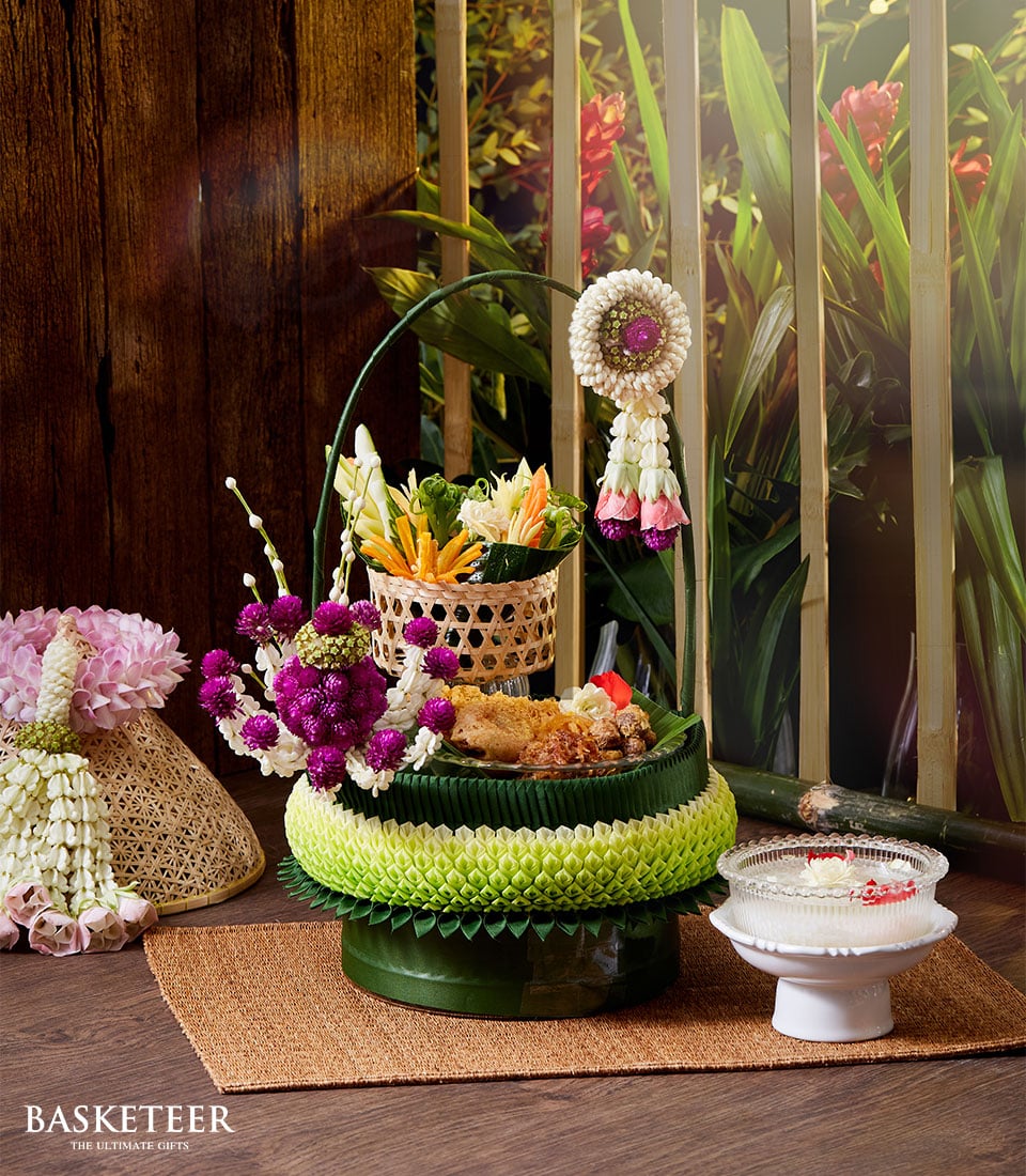 A luxurious decorative basket from Basketeer, evocative of a lavish Songkran celebration, brimming with an exquisite selection of gourmet foods and adorned with vibrant, intricate floral arrangements. This opulent creation is elegantly displayed on a woven mat amidst a rustic yet refined setting, surrounded by potted plants that enhance the inviting ambiance. A white ornate bowl sits gracefully beside an artisanal incense set. The prestigious 