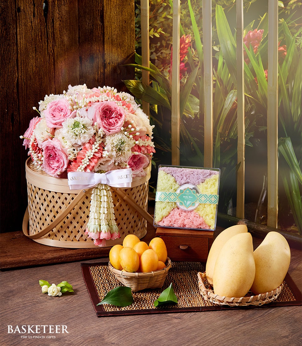 A luxurious wicker basket brimming with delicate pink and white flowers graces a polished table, exuding elegance and charm. Nestled beside it is an exquisitely packaged sweet snack, accompanied by a petite basket of vibrant yellow fruits and two sumptuous golden mangoes. Completing this opulent ensemble is a sophisticated Songkran Thai incense set. The entire scene is framed by rich wooden slats and lush green foliage, enhancing the serene ambiance. A refined label proudly displays the distinguished name 