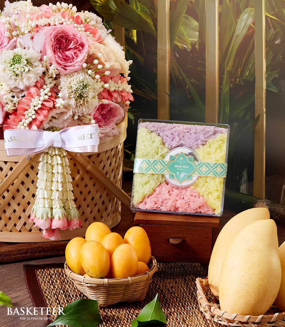 A luxurious wicker basket, meticulously filled with blush pink and pristine white roses, elegantly adorned with a delicate white and pink Basketeer ribbon, rests gracefully on a polished wooden table. Adjacent to it, a charming basket brims with vibrant yellow fruits alongside two perfectly ripe mangoes. A clear box showcases an assortment of pastel-colored treasures from Basketeer. The lush green foliage in the background adds to the tranquil elegance of the scene, evoking the essence of a refined Songkran Thai incense set.