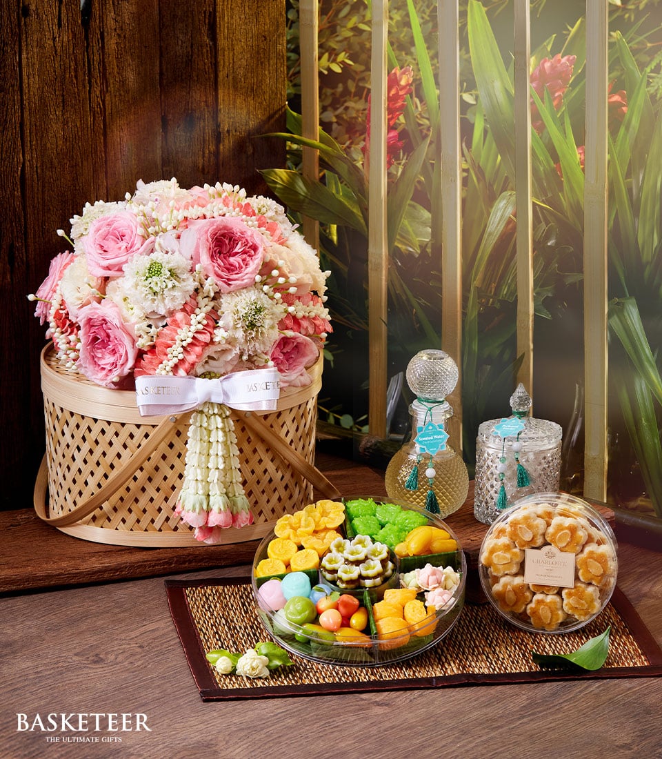An opulent gift set designed for the discerning palate features an elegant basket adorned with lush pink and white floral arrangements, elegantly tied with a pristine white ribbon. A luxurious glass decanter stands alongside a finely decorated container that exudes sophistication. An artisanal wooden board displays an array of vibrant, delectable desserts, inviting indulgence. The prestigious Basketeer branding is subtly positioned at the bottom left, enhancing the aura of exclusivity. This exquisite ensemble is perfect for celebrating Songkran in style, complete with aromatic Thai incense to elevate the experience further.