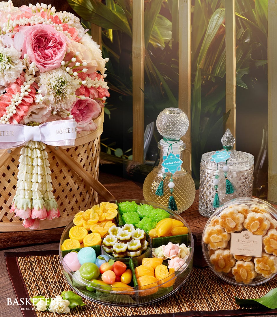 A luxuriously arranged bamboo basket overflowing with elegant pink and white flowers is complemented by two intricately designed bottles from a traditional Songkran Thai incense set, alongside a charming jar. A finely woven mat hosts this lavish display, with an exquisite platter of assorted, brightly colored sweets and a opulent container of gourmet cookies. The scene exudes sophistication and warmth, perfect for any celebration or centerpiece. Look closely and you’ll spot the distinguished Basketeer logo on the delicate ribbon adorning the basket, adding an extra touch of elegance to this already stunning arrangement.