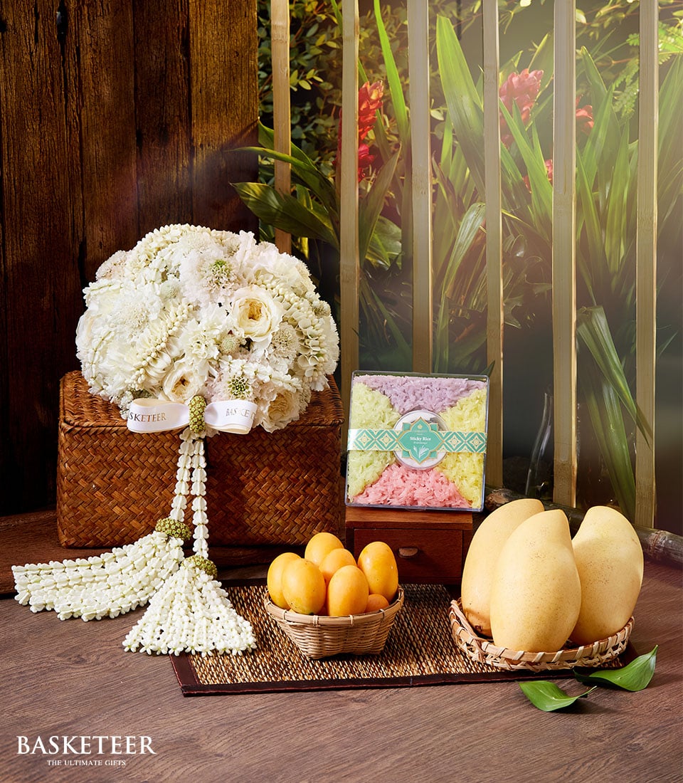 An elegant wicker basket brimming with pristine white flowers and adorned with delicate white floral garlands takes center stage on a luxurious table. Accompanying this exquisite arrangement are three ripe, golden mangoes, a charming round wicker tray overflowing with vibrant small orange fruits, and an artisanal box of colorful treats. The display is beautifully completed by an opulent Songkran Thai incense set. The prestigious 
