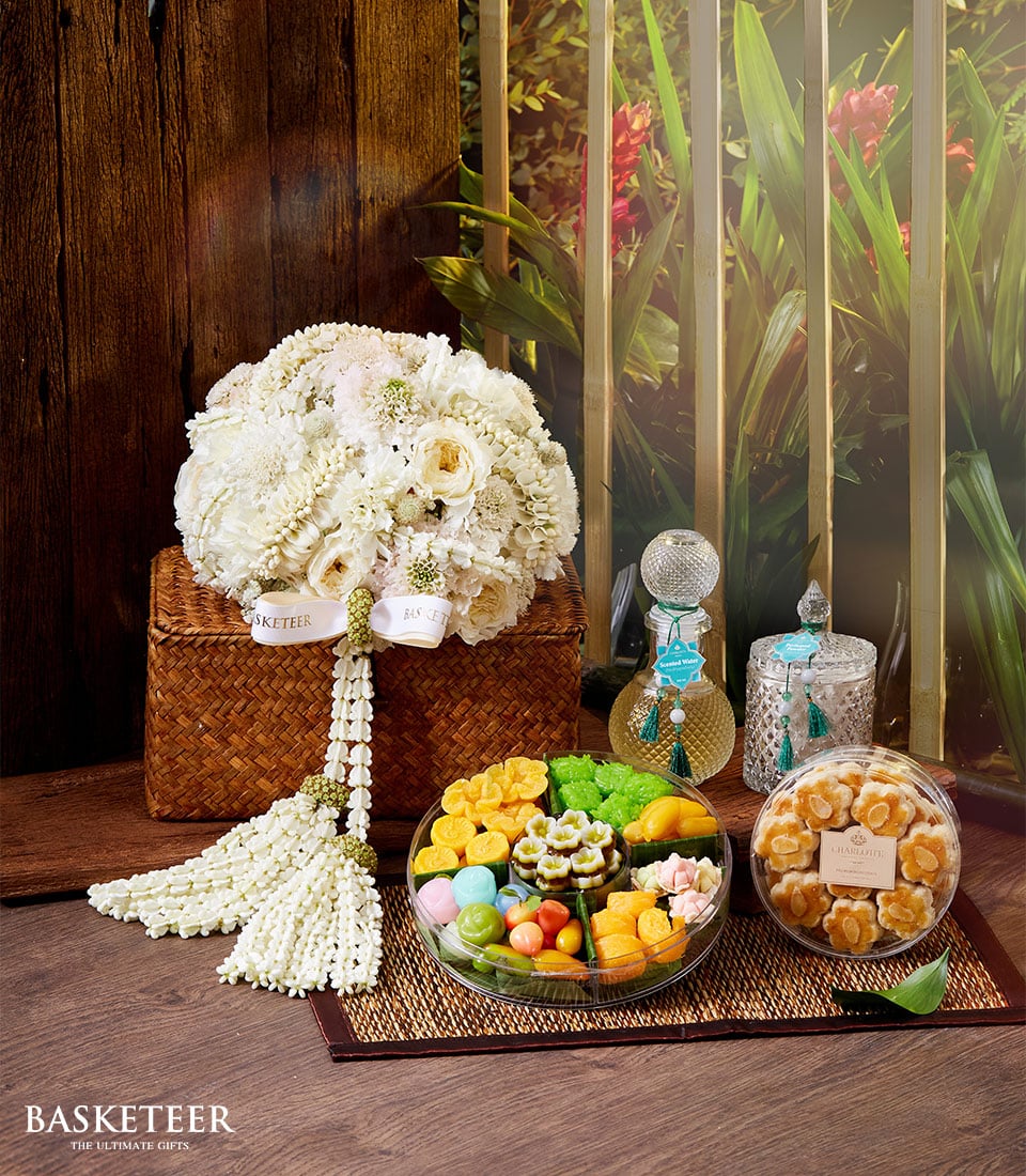 A lavish Basketeer basket brimming with an elegant white floral arrangement and a selection of exquisite traditional Thai desserts. Alongside, a luxurious Songkran Thai incense set, all meticulously presented on a rich wooden floor accented by lush green foliage in the background. The delicacies are thoughtfully arranged on a chic woven tray and housed in pristine clear containers. A ribbon adorning the basket proudly displays the Basketeer logo, underscoring a brand synonymous with luxury and indulgence. 