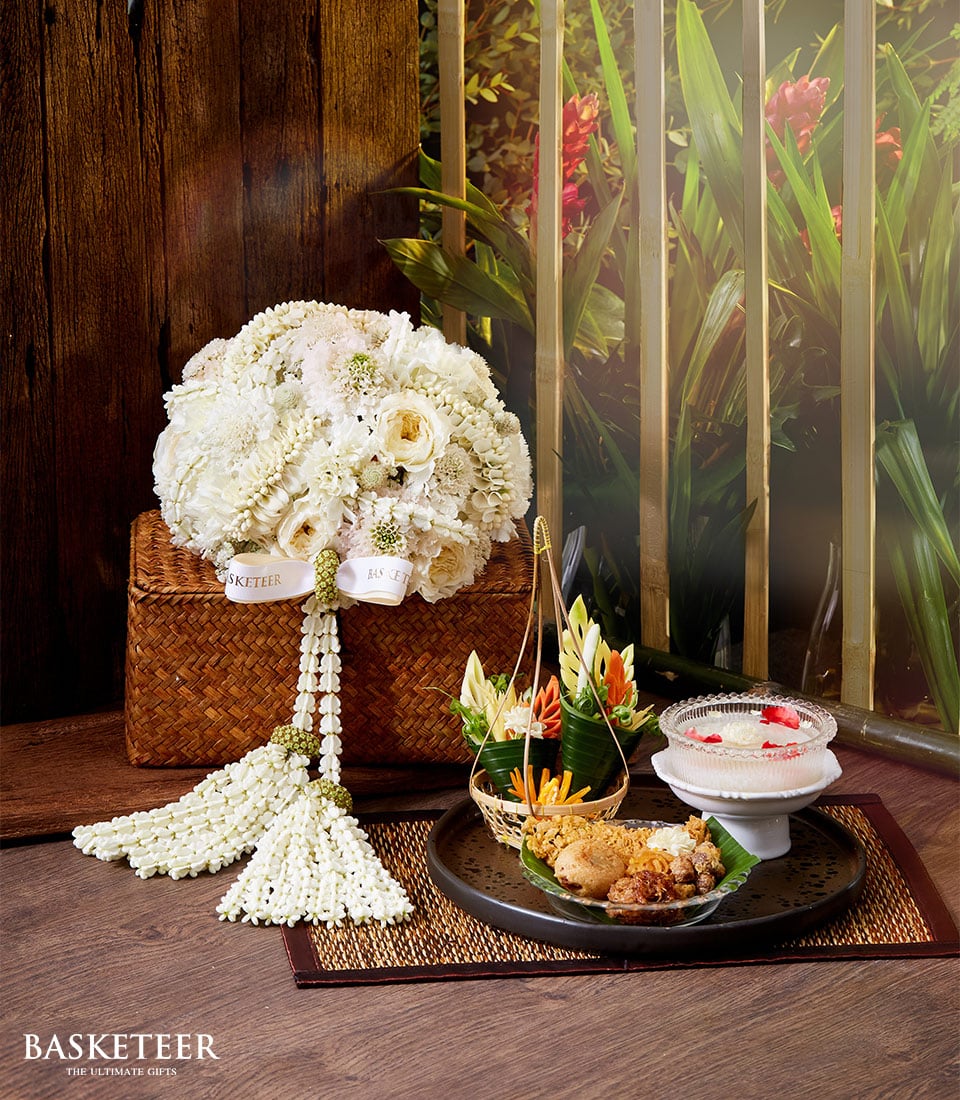 A lavish arrangement showcasing an elegant wicker basket brimming with pristine white flowers, prominently featuring roses. This beautiful setup is paired with an exquisite traditional Thai Songkran meal and an intricate dessert, accompanied by a refined Thai incense set and delicate floral garnishes. The display rests gracefully on a polished wooden surface against the backdrop of a lush, verdant garden. A sophisticated ribbon adorned with the 