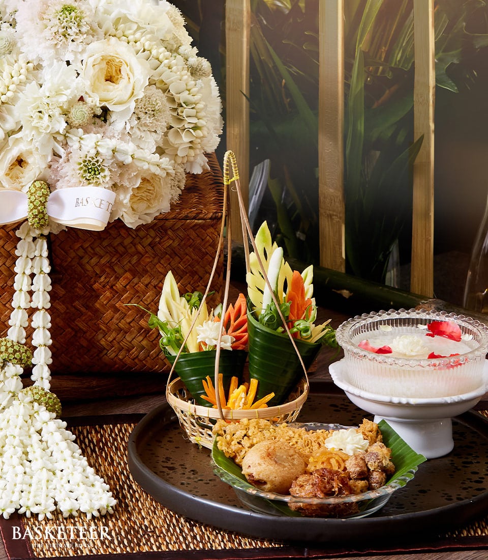 An elegantly curated display showcases a sumptuous bouquet of pristine white flowers intertwined with a lavish garland. A sophisticated basket, adorned with the Basketeer logo, overflows with an array of vividly carved vegetables. A gourmet platter featuring an assortment of exquisitely fried delicacies tempts the senses. Completing this luxurious scene is a glass bowl filled with an indulgent dessert, artfully garnished with delicate rose petals. This traditional Songkran Thai set epitomizes opulence and invites you to indulge in the finest culinary experience by Basketeer.