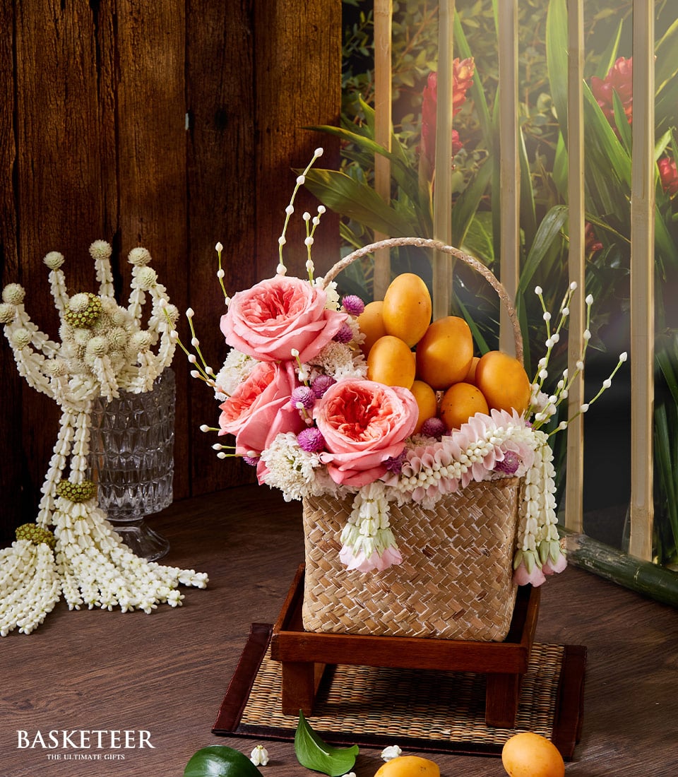 A luxurious woven basket brimming with sun-kissed ripe mangoes is elegantly adorned with delicate pink roses and fragrant jasmine garlands, exuding an enchanting floral perfume. The exquisite basket, featuring the distinguished Basketeer logo, rests gracefully on a refined wooden stand. Additional jasmine garlands cascade over a glass nearby, heightening the sense of opulence. The backdrop of rich wooden panels and lush greenery further amplifies the scene's inviting allure, capturing the essence of Basketeer's timeless elegance.