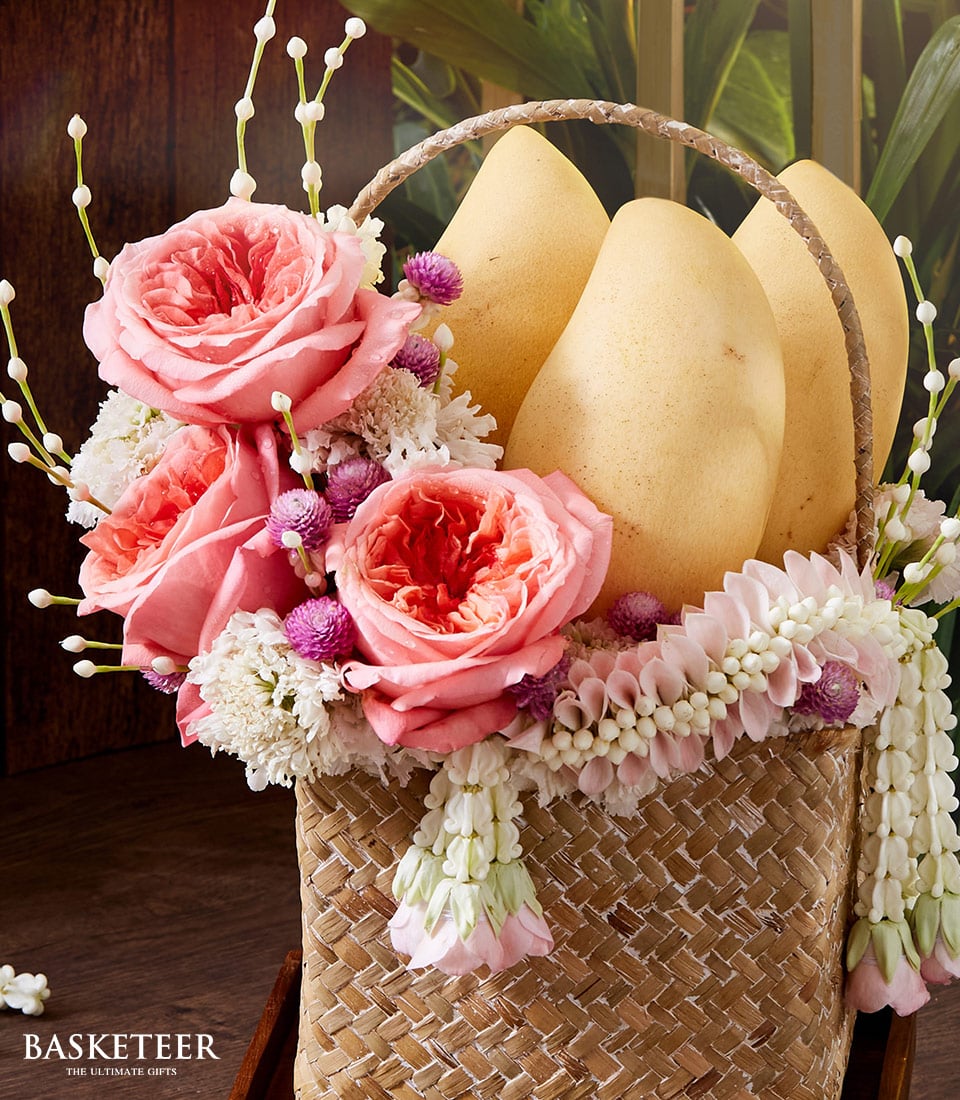 A lavishly woven basket from Basketeer cradles three sun-kissed yellow mangoes, nestled amidst an opulent array of flowers. Radiant pink roses and delicate white blossoms are complemented by a scattering of charming white and purple blooms. This exquisite arrangement embodies luxury and sophistication, perfect for indulging the senses. The serene scene is gracefully completed with the prestigious 