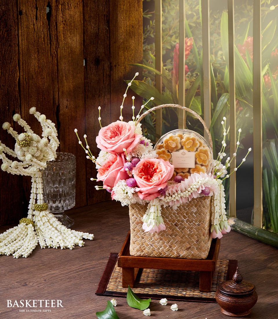 An exquisitely woven basket overflowing with lush pink roses, elegant white blossoms, and intricate Thai flowers graces a polished wooden platform. Beside it, a luxurious glass container is brimming with gourmet almond cookies, tempting the senses. The serene backdrop of rich wooden panels and verdant greenery enhances this opulent setup. A touch of sophistication is added by the Basketeer logo displayed gracefully at the bottom left, signifying unparalleled quality and refinement.