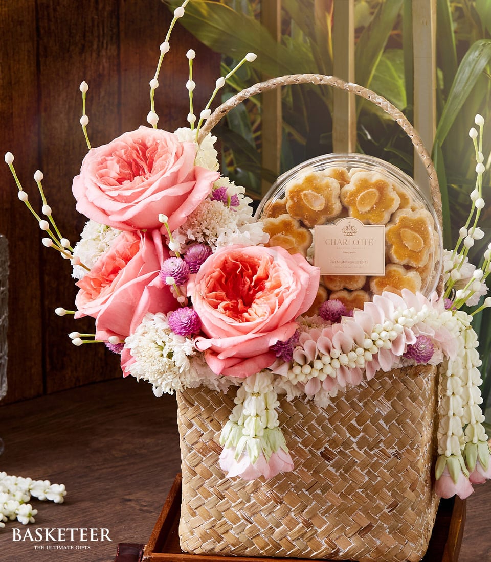 A luxurious woven basket brimming with vibrant pink roses, delicate white and pink blossoms, and accompanied by an elegant round box of gourmet almond cookies. The sophisticated arrangement rests gracefully on a polished wooden surface, framed by lush green foliage that enhances the scene's natural beauty. A distinguished ribbon featuring the 