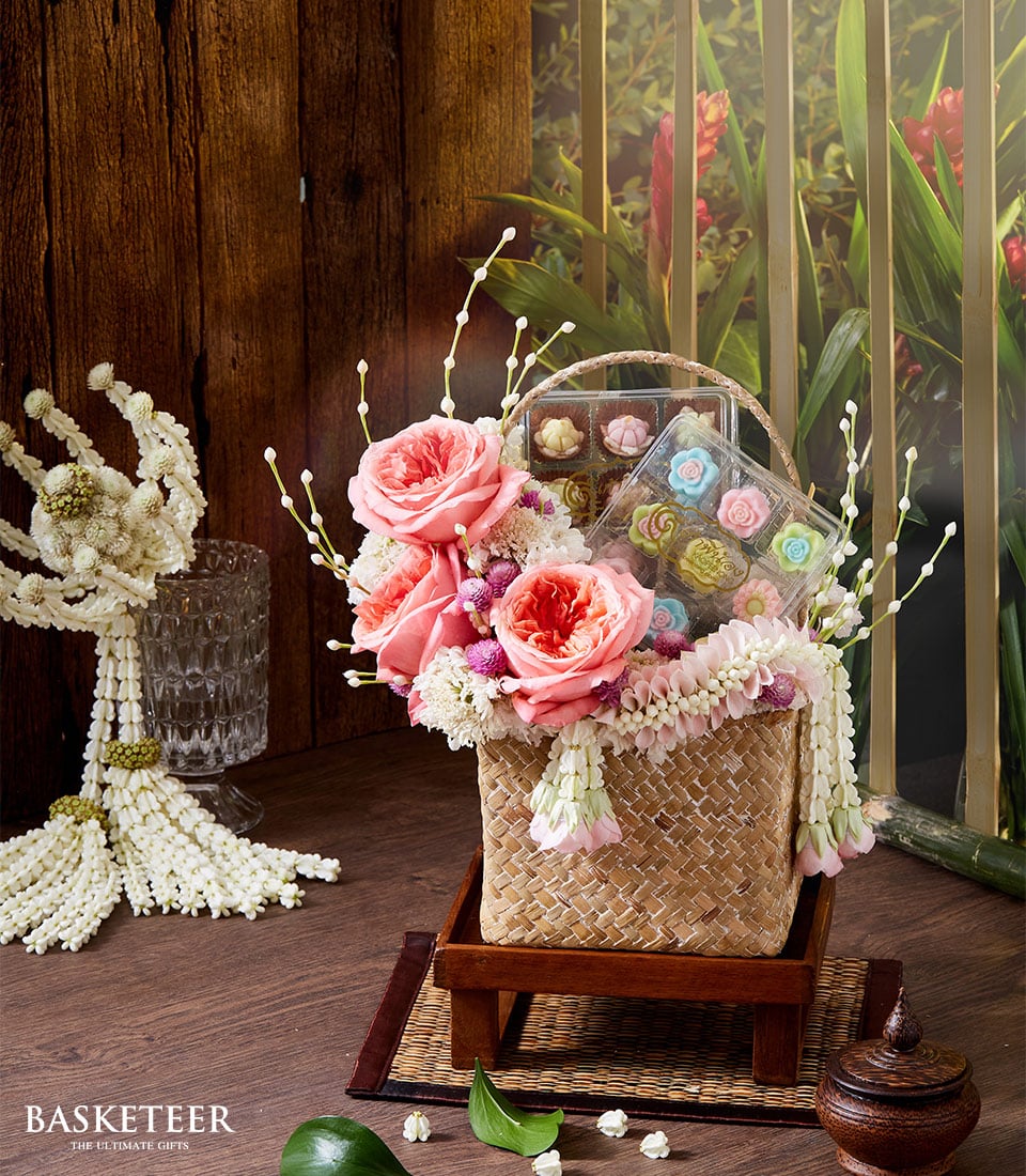 A luxurious wicker basket, elegantly embellished with a cascade of pink roses, delicate white flowers, and an enchanting floral garland. Positioned thoughtfully on an exquisite wooden tray, this opulent offering is accompanied by a clear box showcasing an array of vibrantly colored flower-shaped cookies. The backdrop features rustic wooden walls and a window that frames lush green foliage interspersed with vibrant red blooms. A ribbon elegantly displays the 