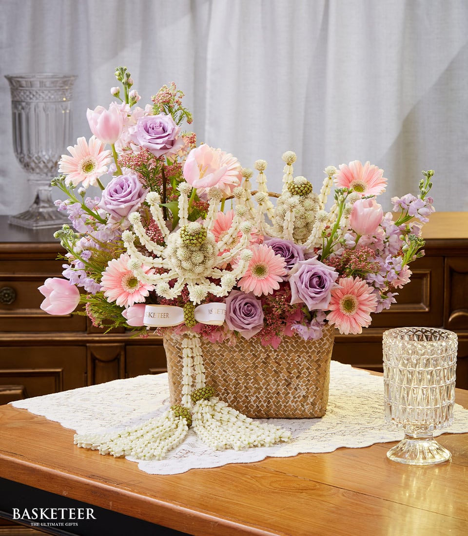 A lavish floral arrangement from Basketeer graces a wooden table adorned with an elegant white lace tablecloth. The woven basket, branded with the distinguished Basketeer emblem, overflows with vibrant pink gerbera daisies, exquisite purple roses, delicate pink tulips, and pristine white decorative accents. Nearby, a crystal glass shimmers in the light, capturing the festive spirit of Songkran. In the background, an ornate cabinet adds to the sophisticated ambiance. Basketeer ensures every detail exudes luxury and charm.