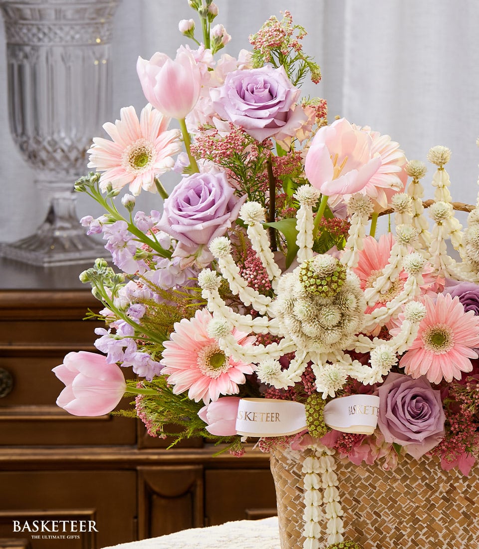 A luxurious woven basket overflowing with an opulent arrangement of pink tulips, delicate pink gerberas, soft light purple roses, and pristine white floral accents. The elegant basket proudly displays the 