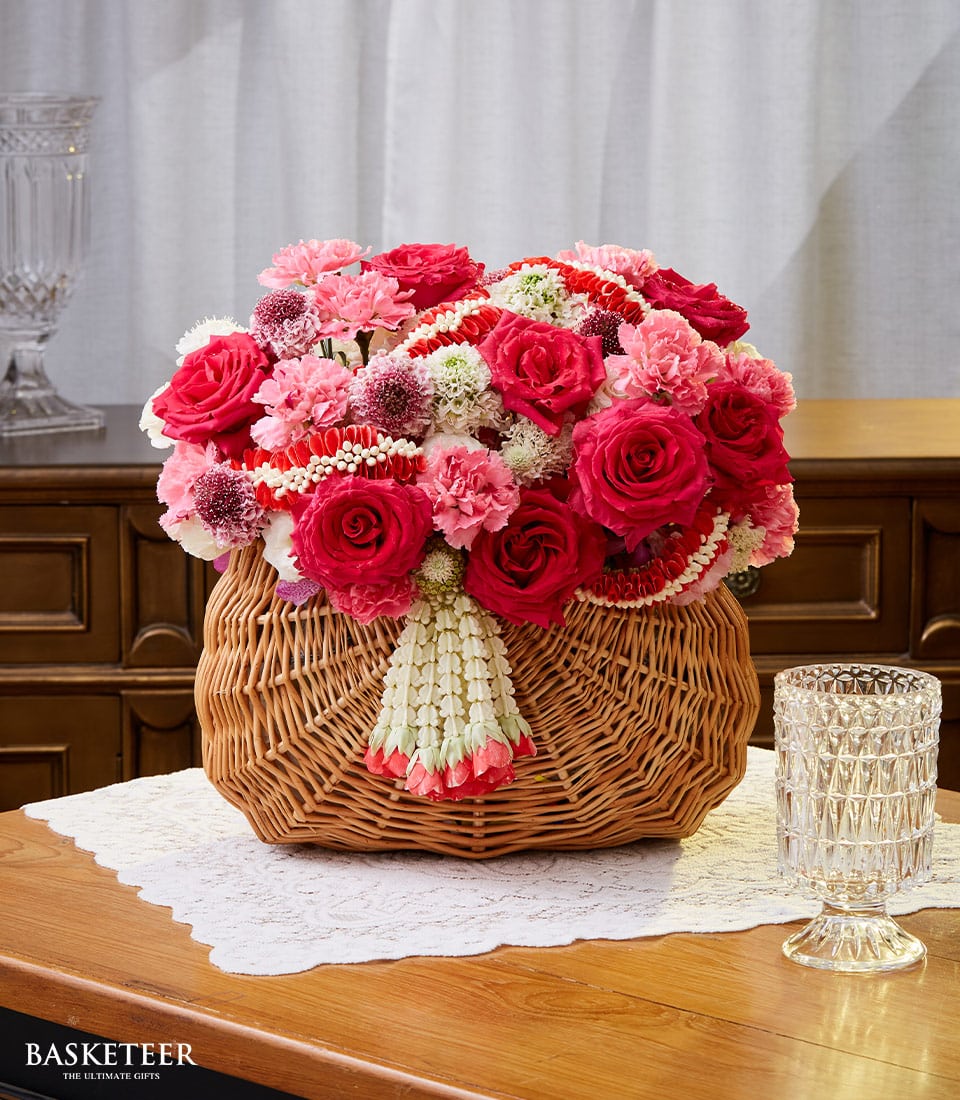 An exquisitely crafted wicker basket from Basketeer, elegantly brimming with a luxurious arrangement of vibrant red roses, delicate pink carnations, and pristine white accents, is gracefully seated atop a finely detailed lace doily on a polished wooden table. The basket's charm is further enhanced by an artfully draped floral garland reminiscent of opulent Songkran festivities. Beside the ornate display sits crystal glassware that adds a touch of sophistication to the scene. Sheer curtains in the background gently filter natural light, casting an inviting and serene ambiance over this lavish setting. Look closely to find the Basketeer logo subtly gracing a ribbon or box within this beautifully composed tableau.