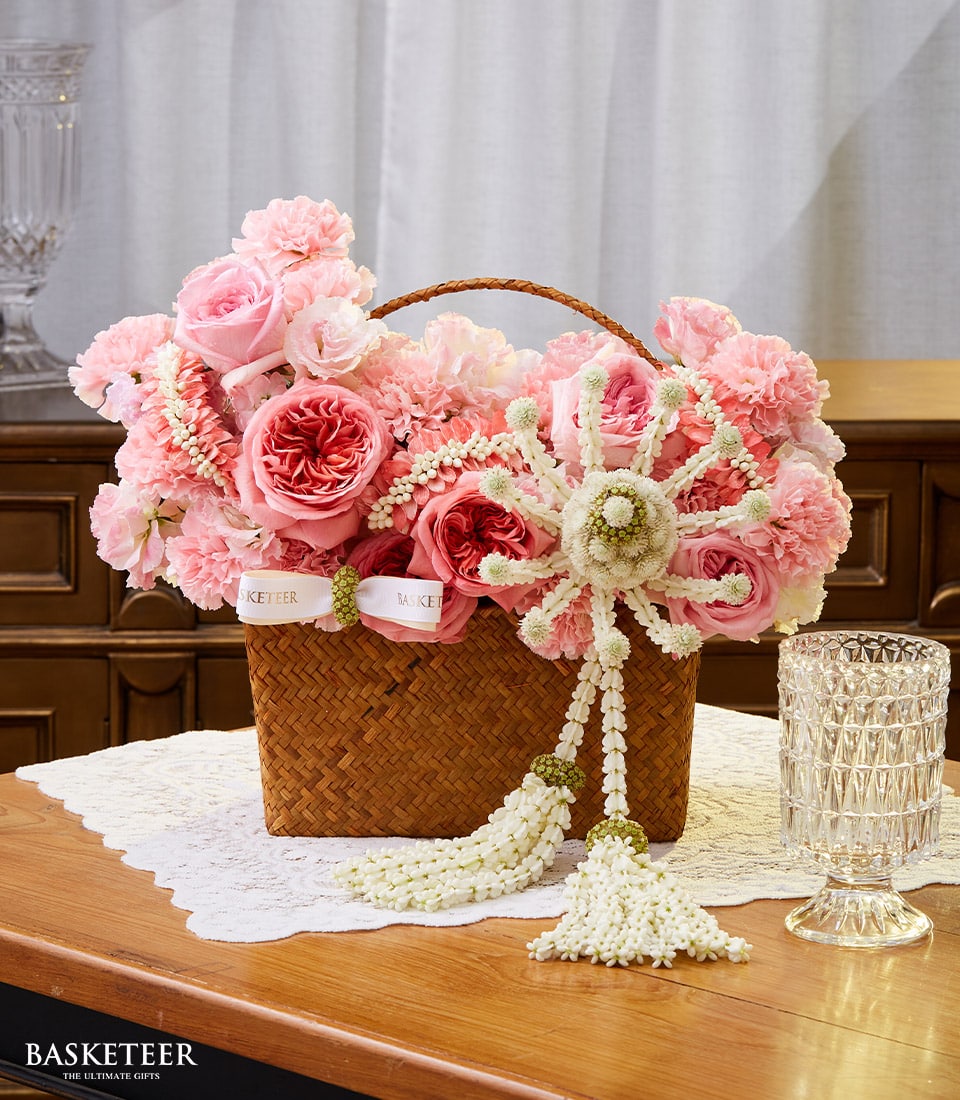 A lavish wicker basket, elegantly filled with a stunning floral arrangement of pink roses and carnations from Basketeer, graces a lace doily atop a polished wooden table. A delicate white floral garland weaves through the blossoms, exuding the essence of Songkran's enchanting beauty. Nearby, an ornate glass adds a touch of sophistication to the scene. The backdrop features an exquisite wood-paneled chest and soft-flowing white curtains, completing this picture of refined luxury by Basketeer.