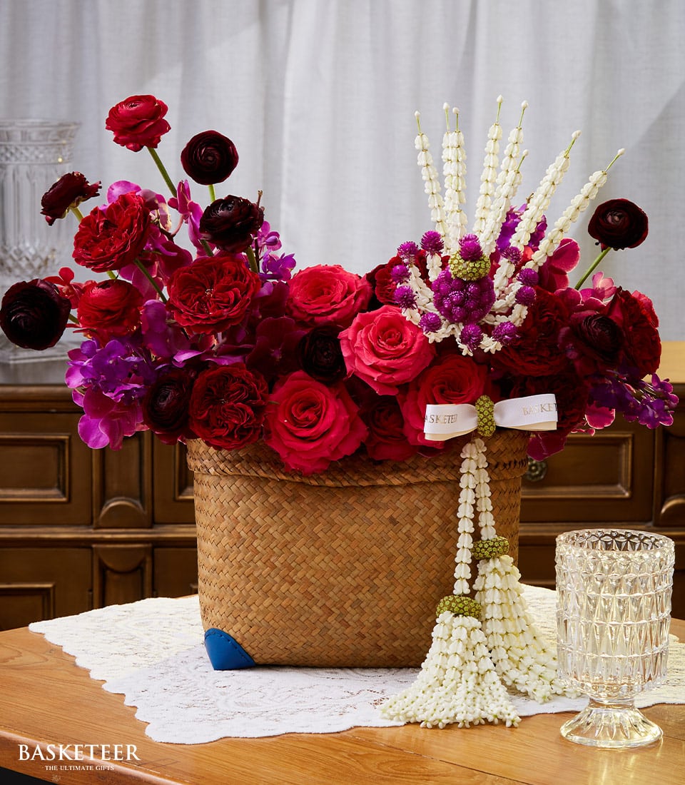 A luxurious woven basket, carefully curated by Basketeer, brims with a stunning floral arrangement featuring an opulent blend of red and pink roses, deep purple ranunculus, and bright pink orchids. Draped elegantly over the basket is a traditional Thai flower garland crafted from pristine white jasmine, making it an exquisite Songkran gift. Nearby, a crystal glass adds an extra touch of sophistication to the scene. The essence of Basketeer's refinement is palpable in every detail.
