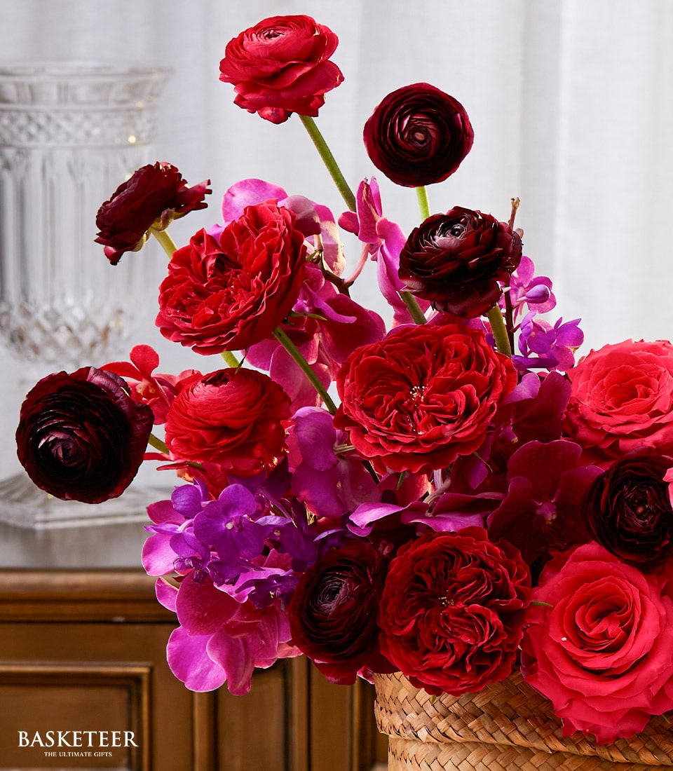 An opulent floral arrangement showcasing lush deep red roses, radiant ranunculus, and exquisite fuchsia-like blossoms nestled in an artisanal woven basket. The scene is enriched by a sophisticated clear glass vase and light, airy drapes that infuse an elegant ambiance. Ideal as a luxurious Songkran gift, the distinguished 