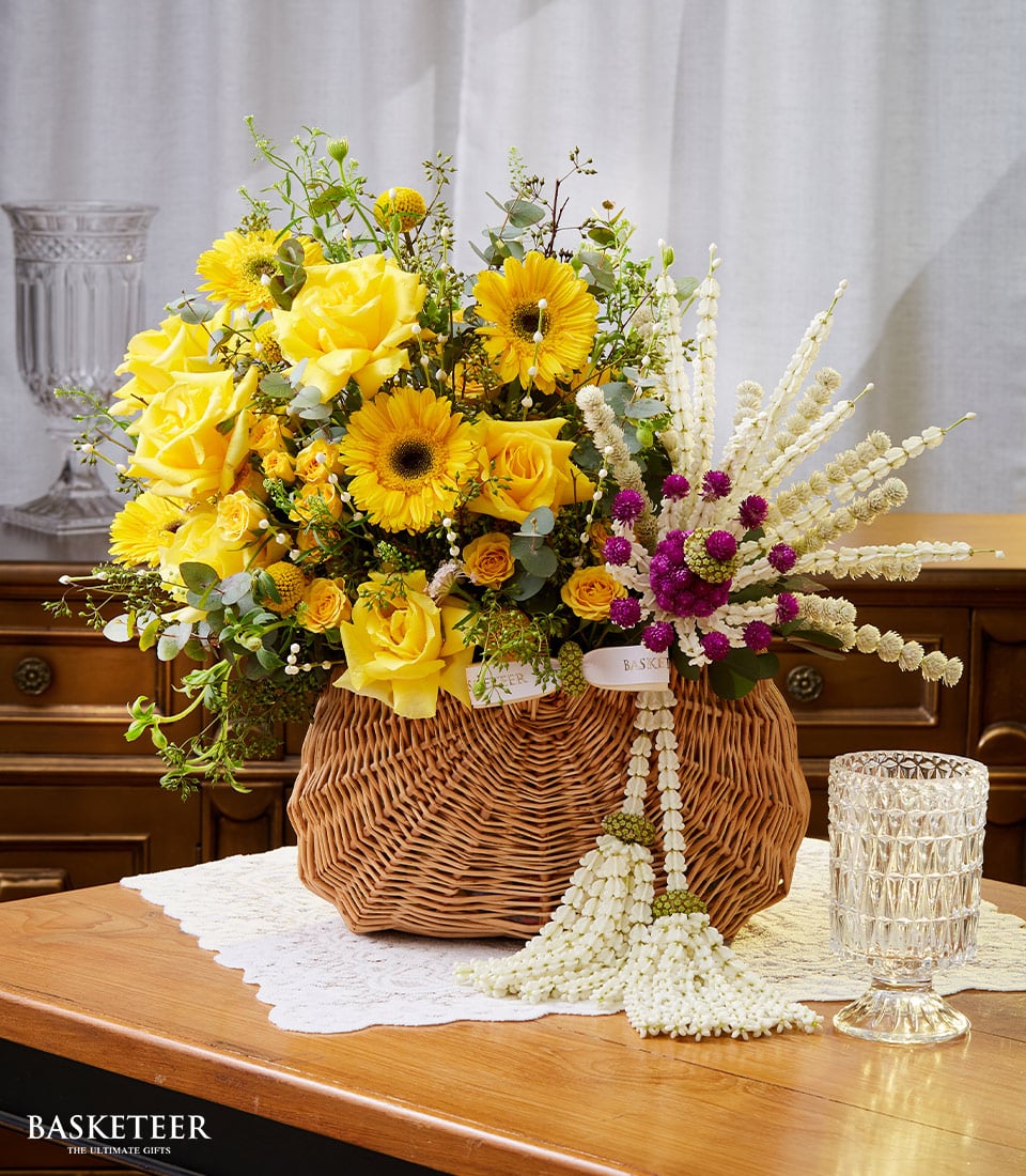 A luxurious wicker basket overflowing with a stunning arrangement of vibrant yellow roses, radiant sunflowers, and lush greenery from Basketeer graces a wooden table adorned with an exquisite white lace cloth. The basketeer-branded ribbon elegantly wraps around the basket. Beside this opulent display lies a delicate Songkran garland interwoven with pristine white and enchanting purple blossoms. In the background, a sparkling crystal glass and an artfully designed vase add to the scene's sophisticated allure.