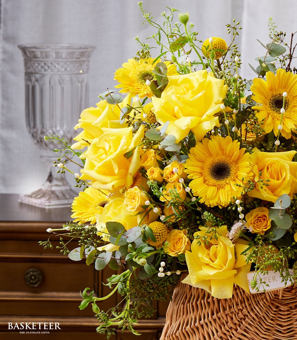 A splendid bouquet of luxurious yellow roses, radiant sunflowers, and lush greenery elegantly arranged in a refined wicker basket. The ensemble is gracefully set atop an exquisite wooden furniture piece, exuding an atmosphere of opulence and warmth. In the backdrop, a meticulously crafted clear ornate vase adds a touch of sophistication on the left side, while light-colored drapery cascades gently behind the arrangement. Ideal for gifting during Songkran, this Basketeer creation promises to elevate any celebration with its breathtaking beauty. The distinguished 