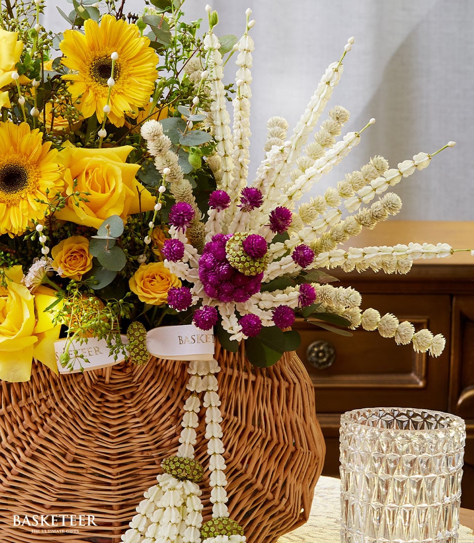 A luxurious wicker basket brimming with radiant yellow sunflowers and vibrant yellow roses, intertwined with lush green foliage, epitomizes the elegance of a Songkran floral gift. The arrangement is graced by delicate cascades of white and purple dried flowers adorning its sides. Resting on a polished table, this exquisite floral masterpiece is complemented by a crystal glass filled with refreshing water. Intricately tied around the basket is a satin ribbon bearing the Basketeer logo, adding an exclusive touch to this inviting scene.