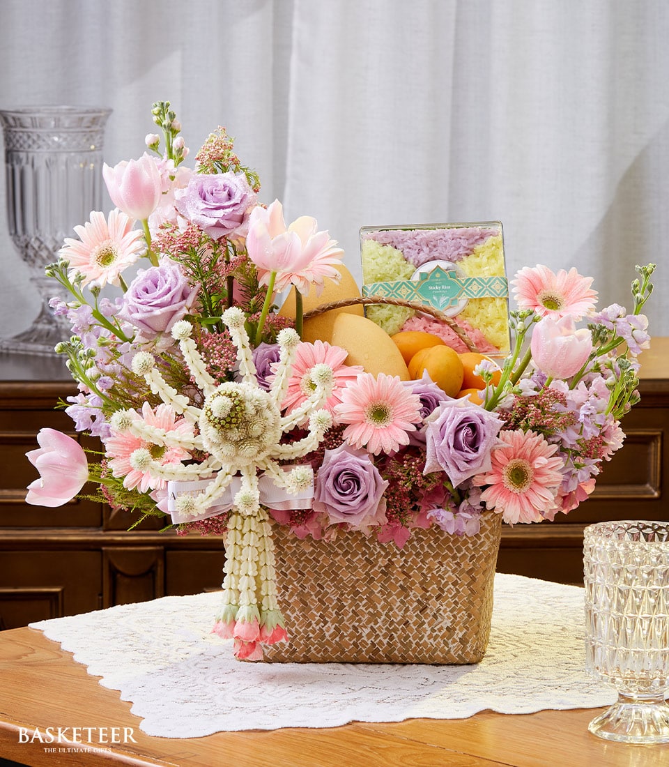 A luxurious woven basket by Basketeer graces the refined tabletop, brimming with a spectacular Songkran floral arrangement. Delicate pink tulips, resplendent purple roses, and cheerful pink daisies nestle together in captivating harmony, exuding an air of opulence. A succulent orange fruit adds a touch of zest to the ensemble, which is further elevated by an elegantly packaged gift adorned with the distinguished Basketeer logo. The backdrop of flowing white curtains and exquisite glassware enhances the sumptuous ambiance, perfectly capturing the essence of festive sophistication.