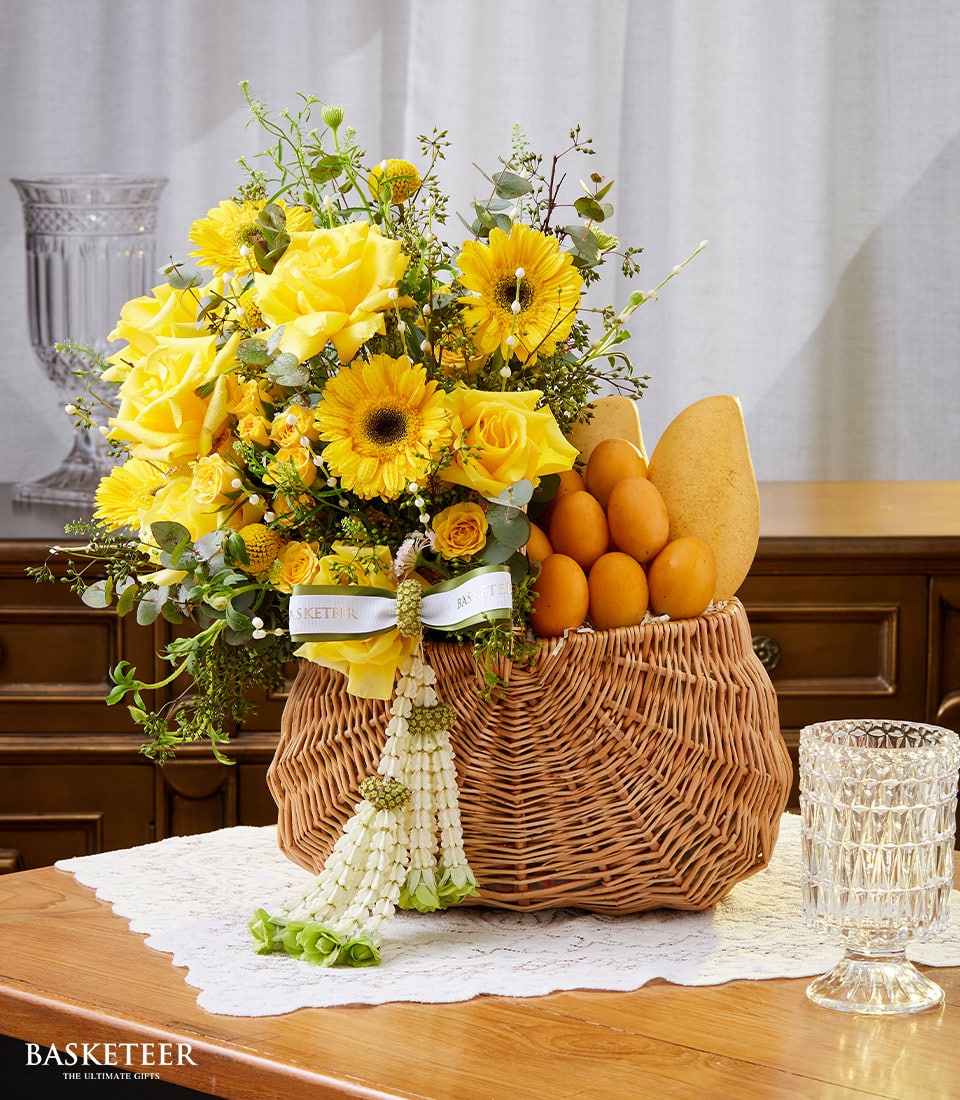 A luxurious wicker basket from Basketeer, elegantly presented on a delicate lace tablecloth, cradles a vibrant arrangement of radiant yellow roses, cheerful sunflowers, and lush green foliage—a true Songkran-inspired masterpiece. Nestled beside the bountiful blooms are an assortment of golden-brown artisan breads. A crystal glass gleams nearby on a polished wooden table, completing the scene with an air of refined sophistication.